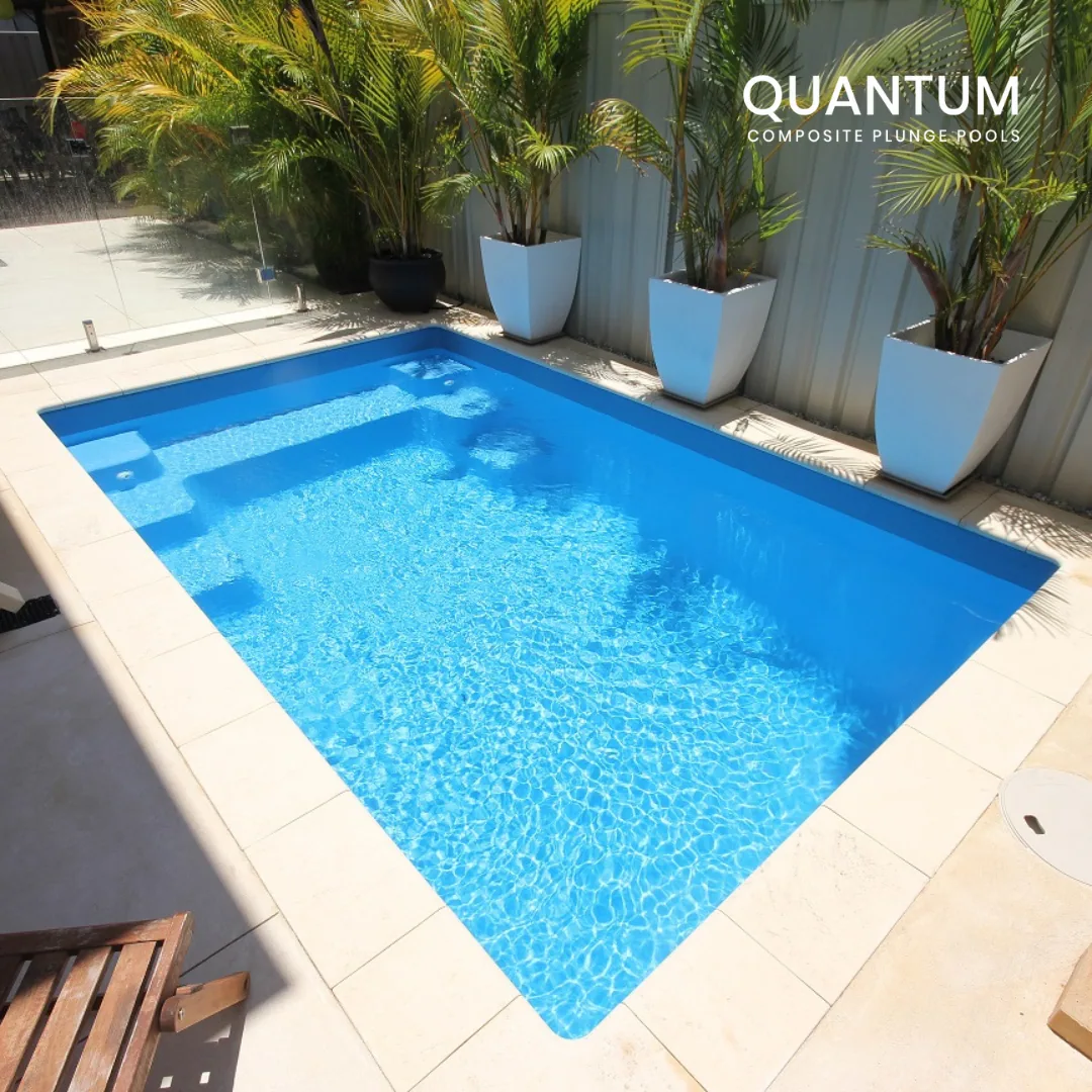 A small rectangular backyard pool with blue water, surrounded by white tile deck and large potted tropical plants, with a metal fence and greenery in the background.