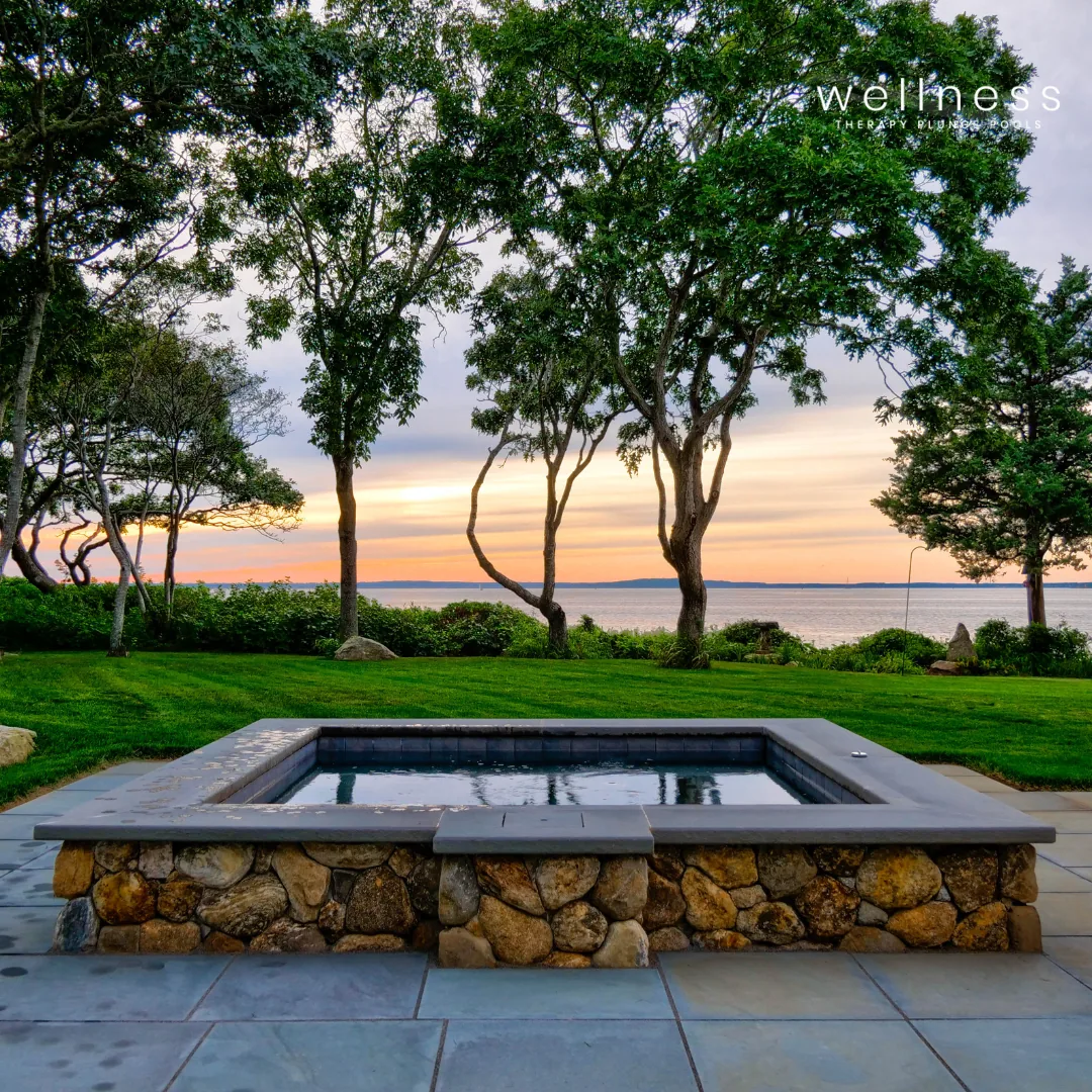 A spa hot tub built with stone and concrete, overlooking a lake during sunset, with trees and grassy area in the background.