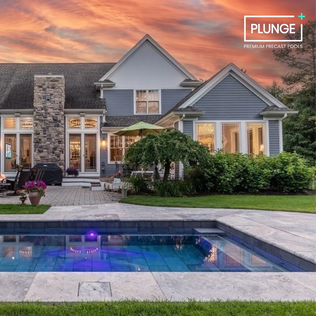 A house with blue siding, stone chimney, and large windows at sunset. There is a backyard with a pool, greenery, and outdoor furniture, including a grill and an umbrella.