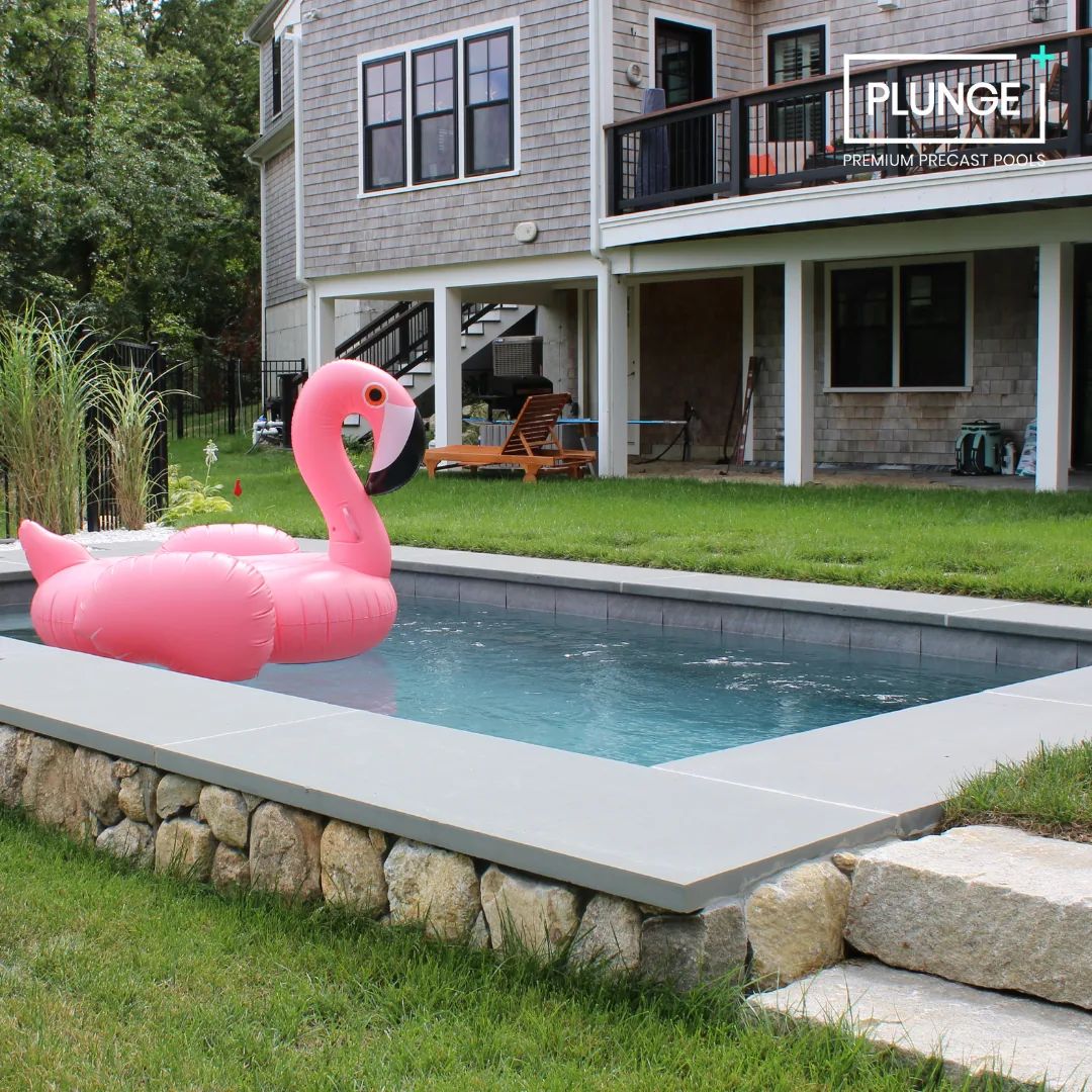 A backyard with a swimming pool held by a stone wall, a pink flamingo pool float, green grass, and a two-story house in the background with a balcony and patio furniture.