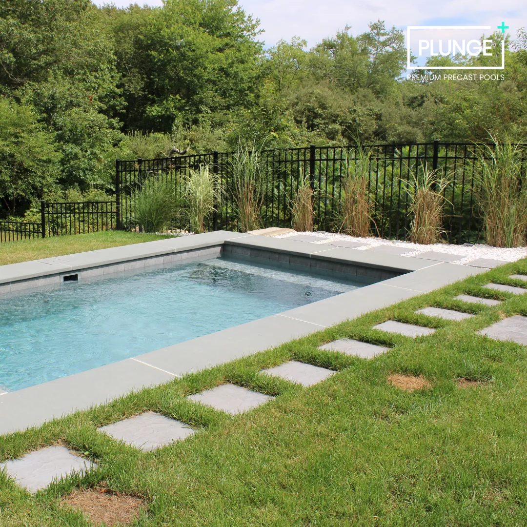 A rectangular backyard pool with concrete edging, surrounded by grass and a stepping stone path, with a black metal fence and green trees in the background.