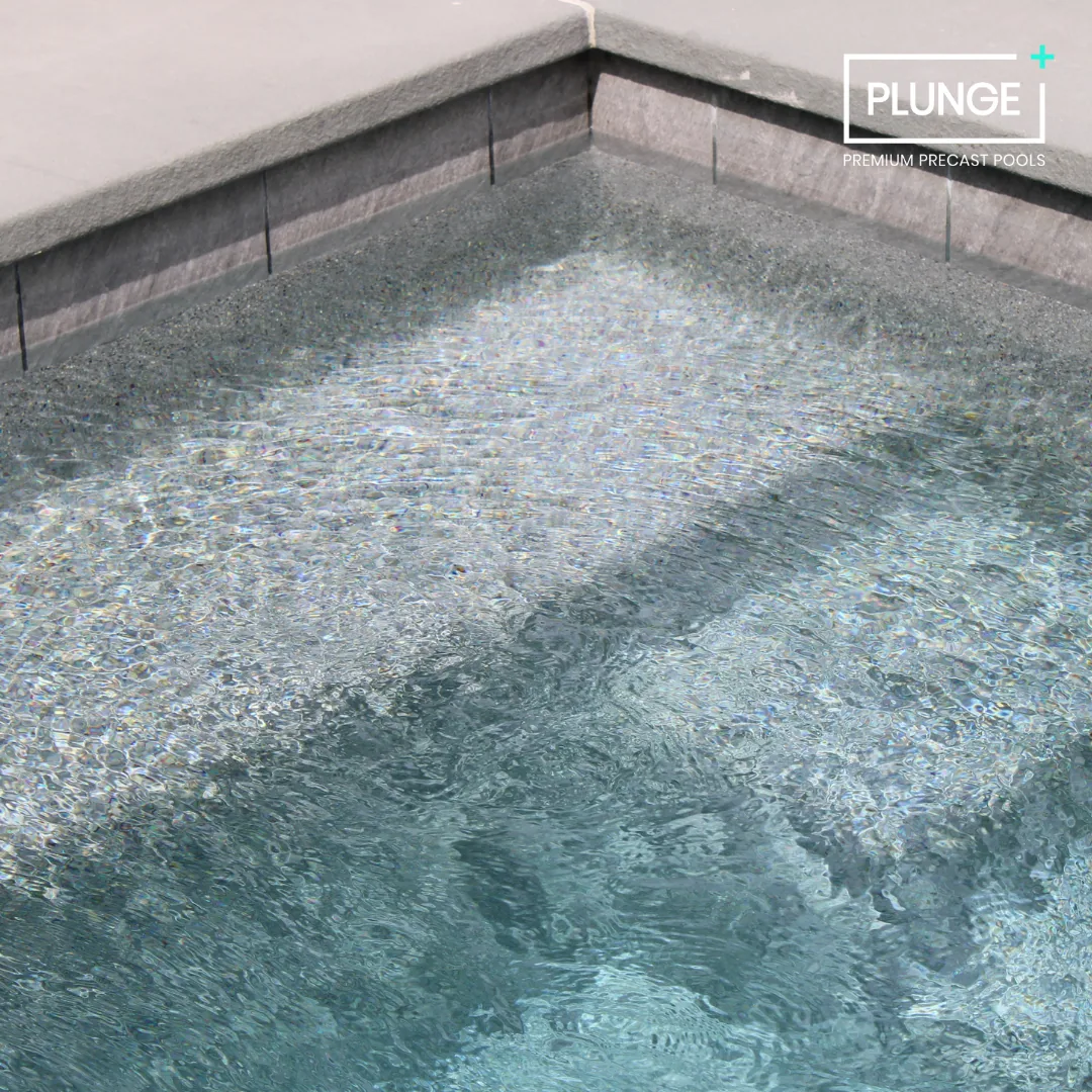 Close-up of a luxury swimming pool with clear water and textured pool edge tiles, with a logo in the corner that reads 'PLUNGE Premium Precast Pools.'