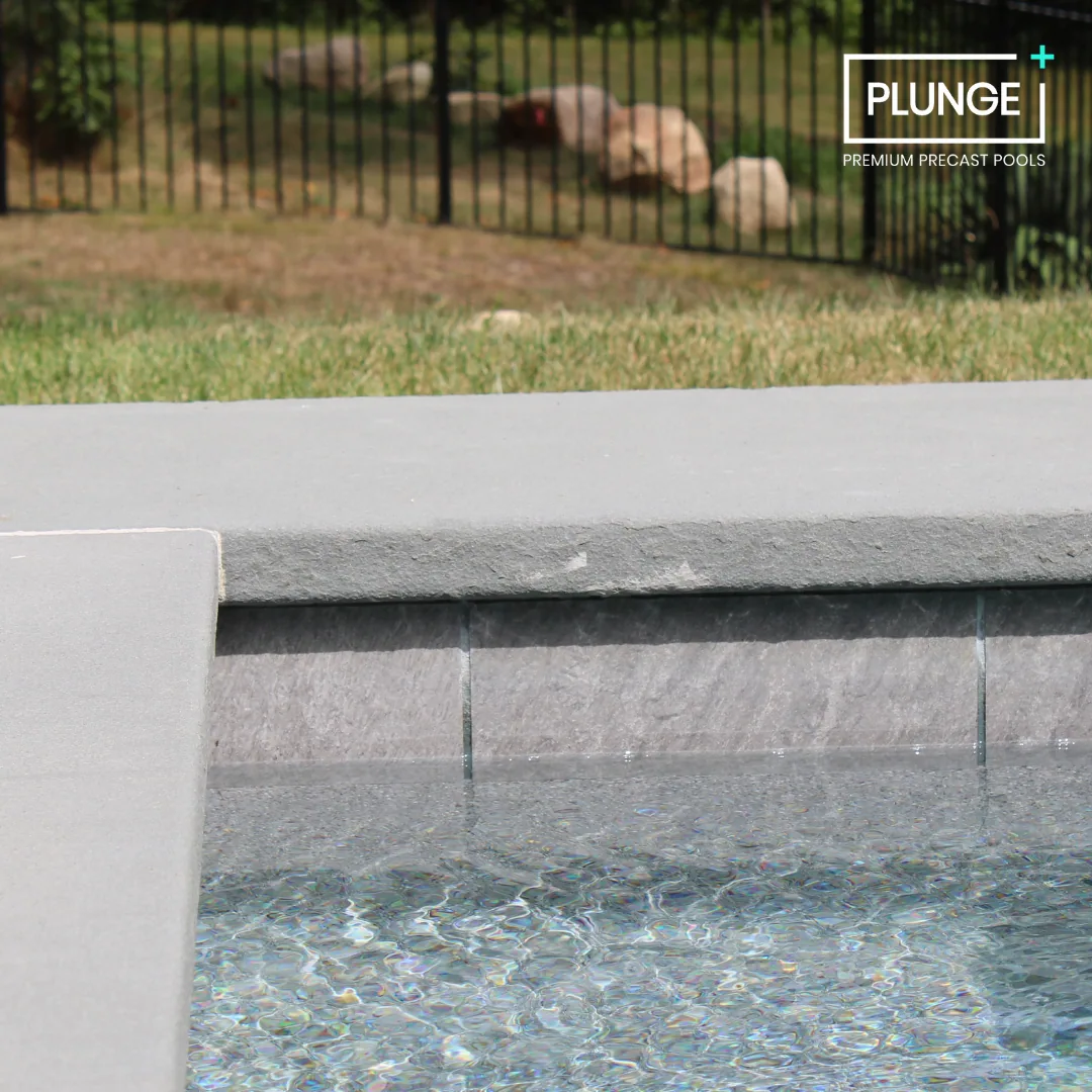 Close-up of a modern concrete pool edge with a water feature, fence, grass, and rocks in the background.