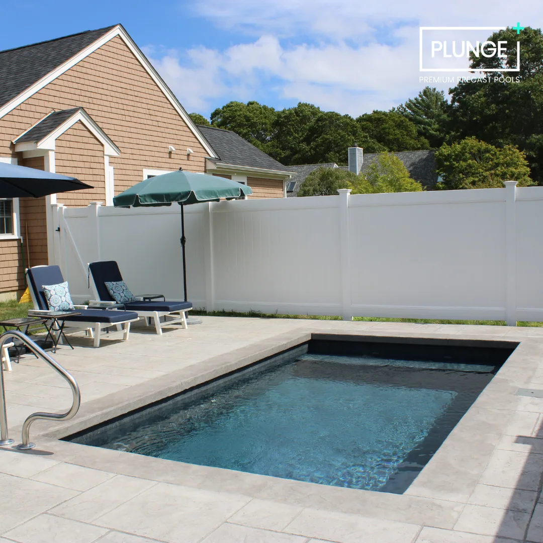 A backyard with a small swimming pool, two lounge chairs with cushions, a patio umbrella, and a white privacy fence. Houses and trees are visible in the background under a partly cloudy sky.
