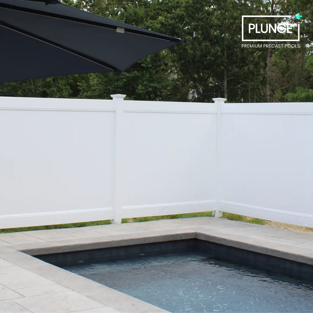 A backyard pool with a white privacy fence, a large dark blue umbrella, and trees in the background. The pool has a stone border and is filled with water.
