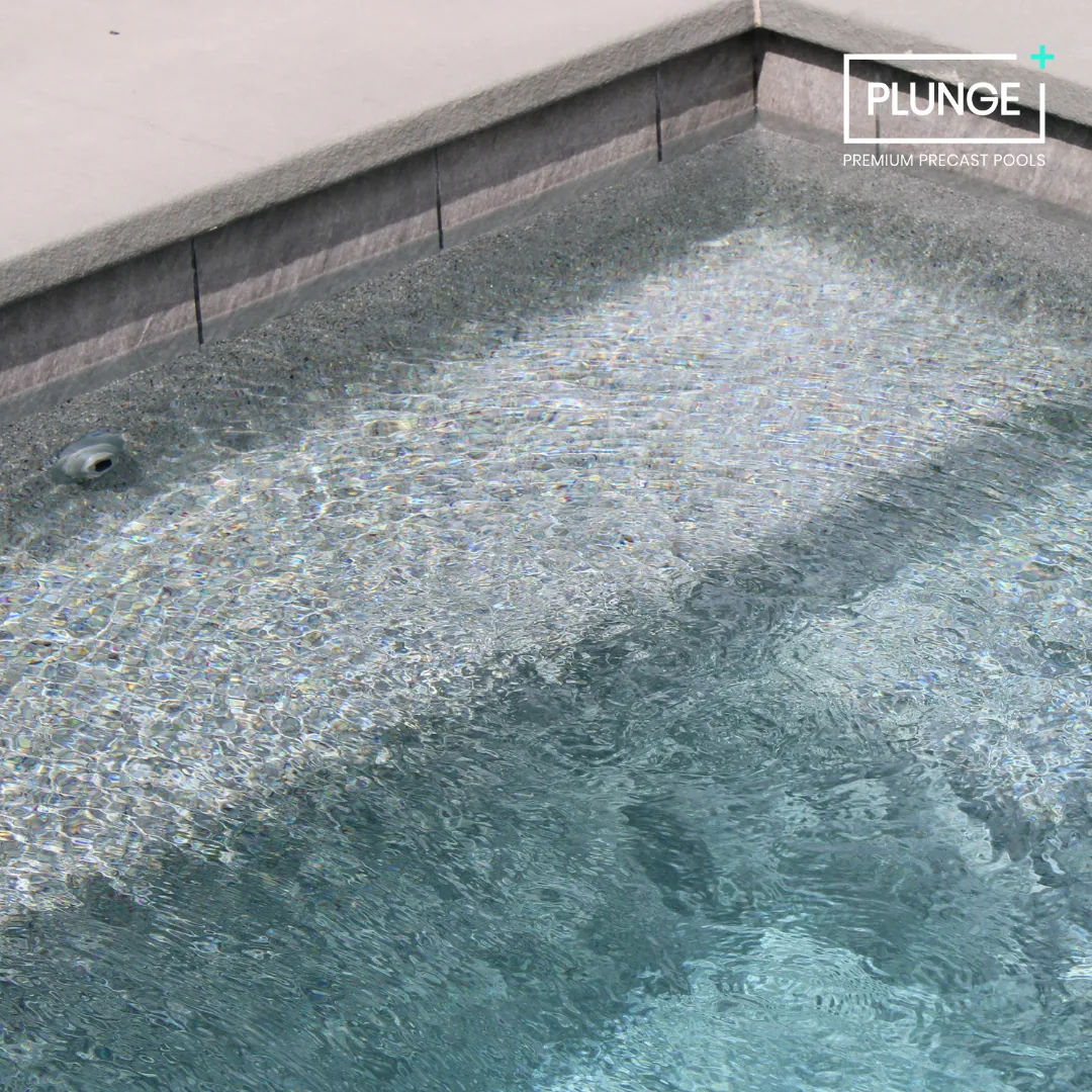 Close-up of a corner of a luxury outdoor inground pool with clear water, tiled pool edge, and a gray concrete deck. The pool has a premium precast concrete pool wall and a water jet fitting.