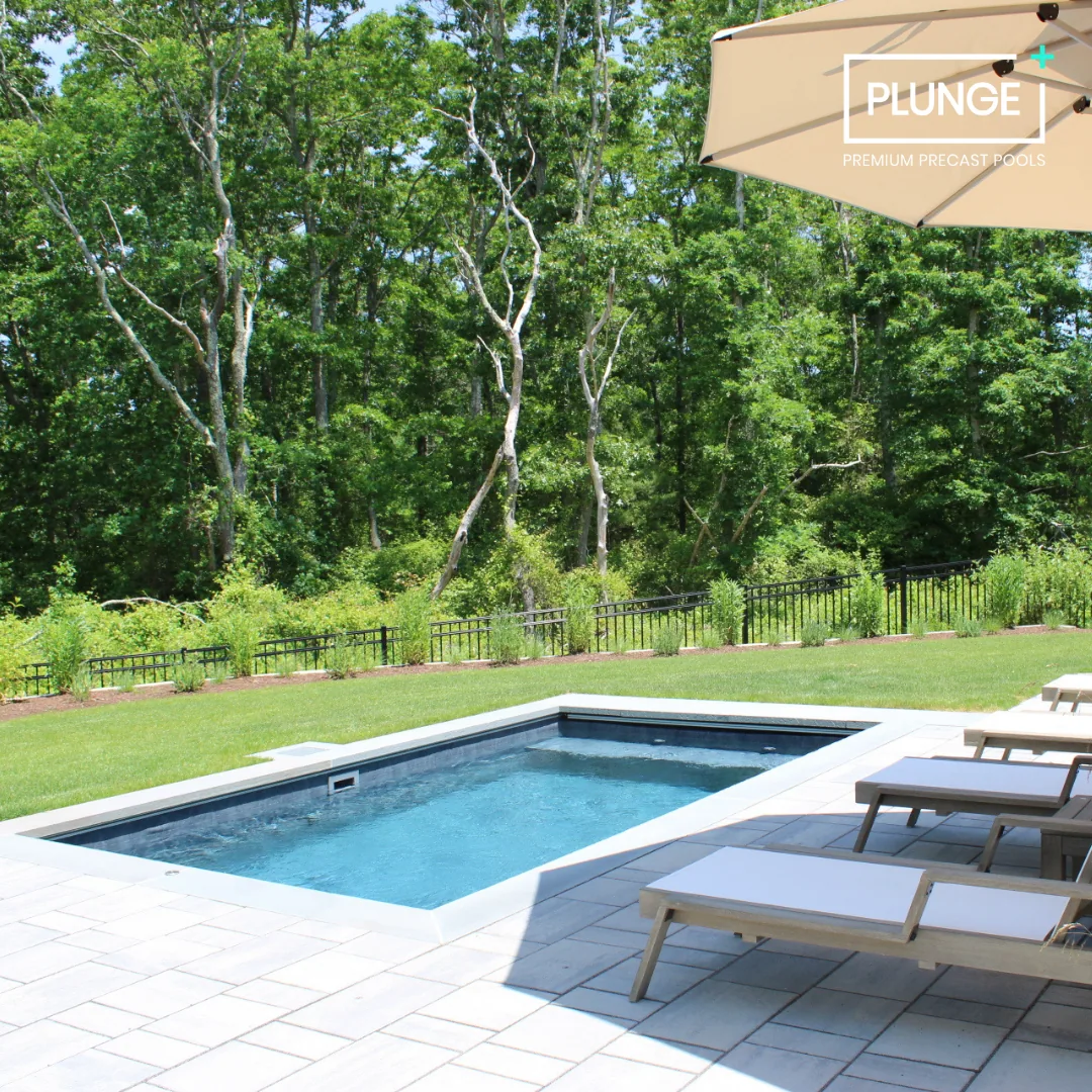 A backyard with a small rectangular pool, sun loungers, an umbrella, green grass, and trees in the background.