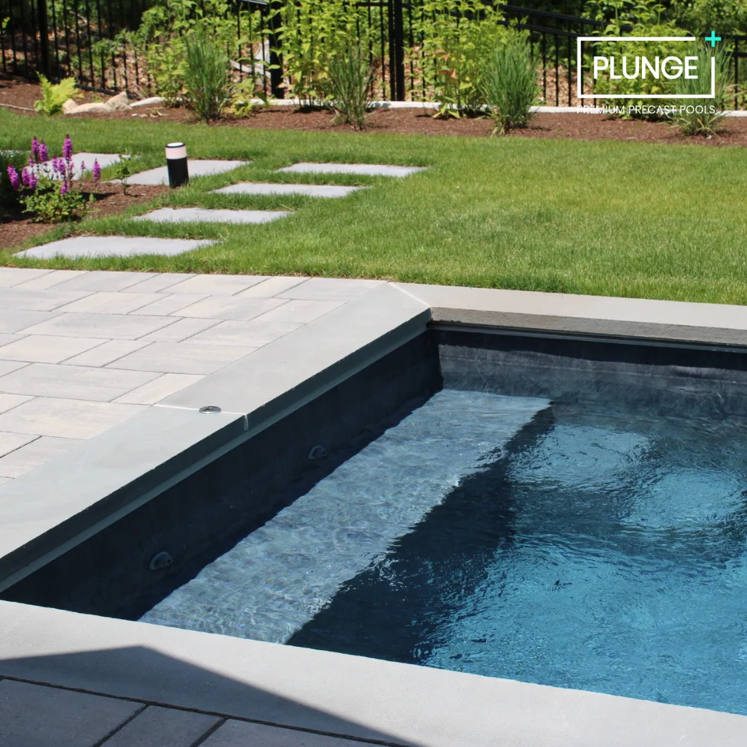 A backyard with green grass, a stone pathway, shrubs, and flowers, next to a black-lined swimming pool with a concrete deck.