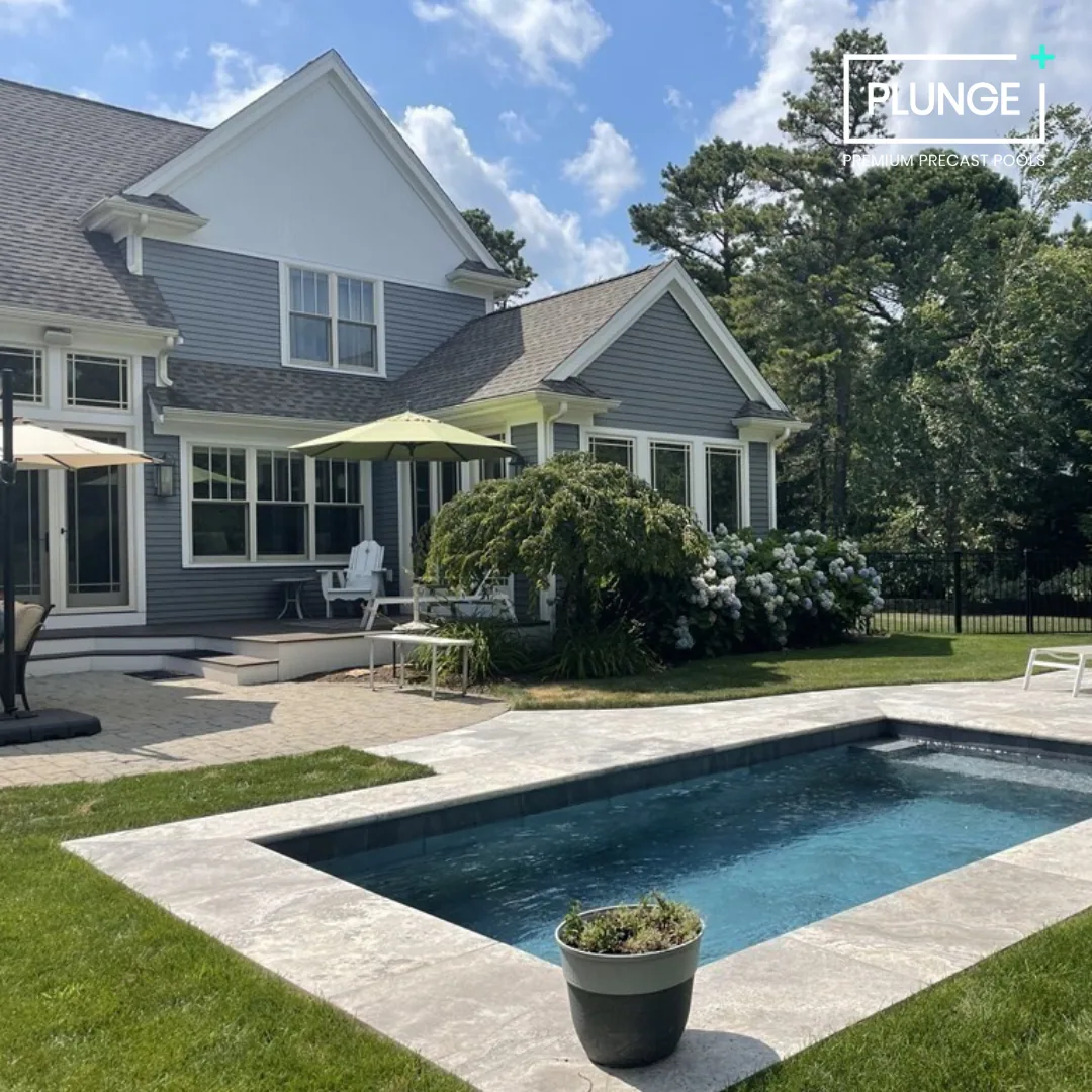 A backyard with a swimming pool, a gray house with large windows, green lawn, and trees in the background. Outdoor patio furniture and umbrellas are visible, along with flowering bushes.