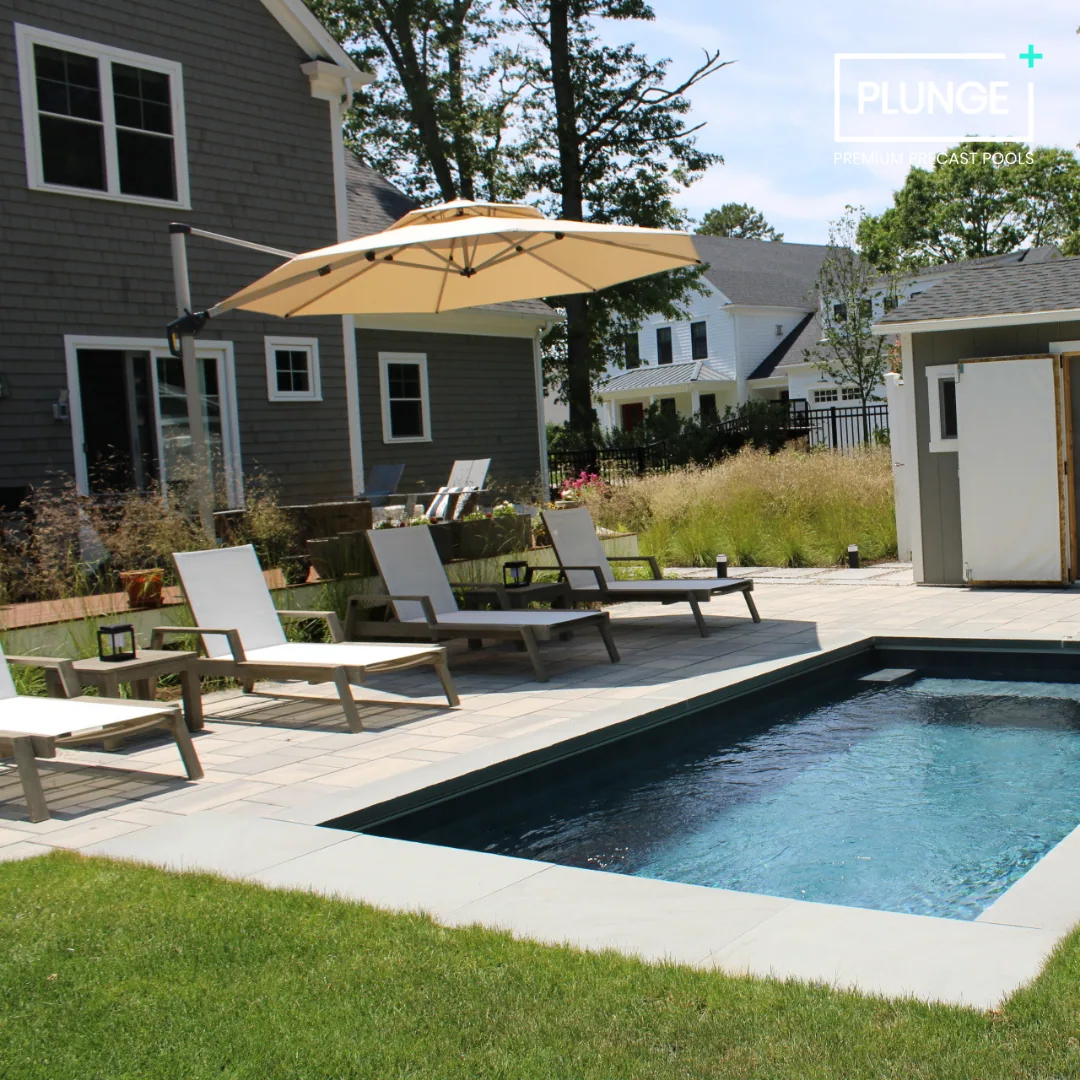 A backyard with a rectangular in-ground swimming pool, several white lounge chairs with black frames, a large beige umbrella, and a shed. There is grass and plants in the background with houses and trees beyond.