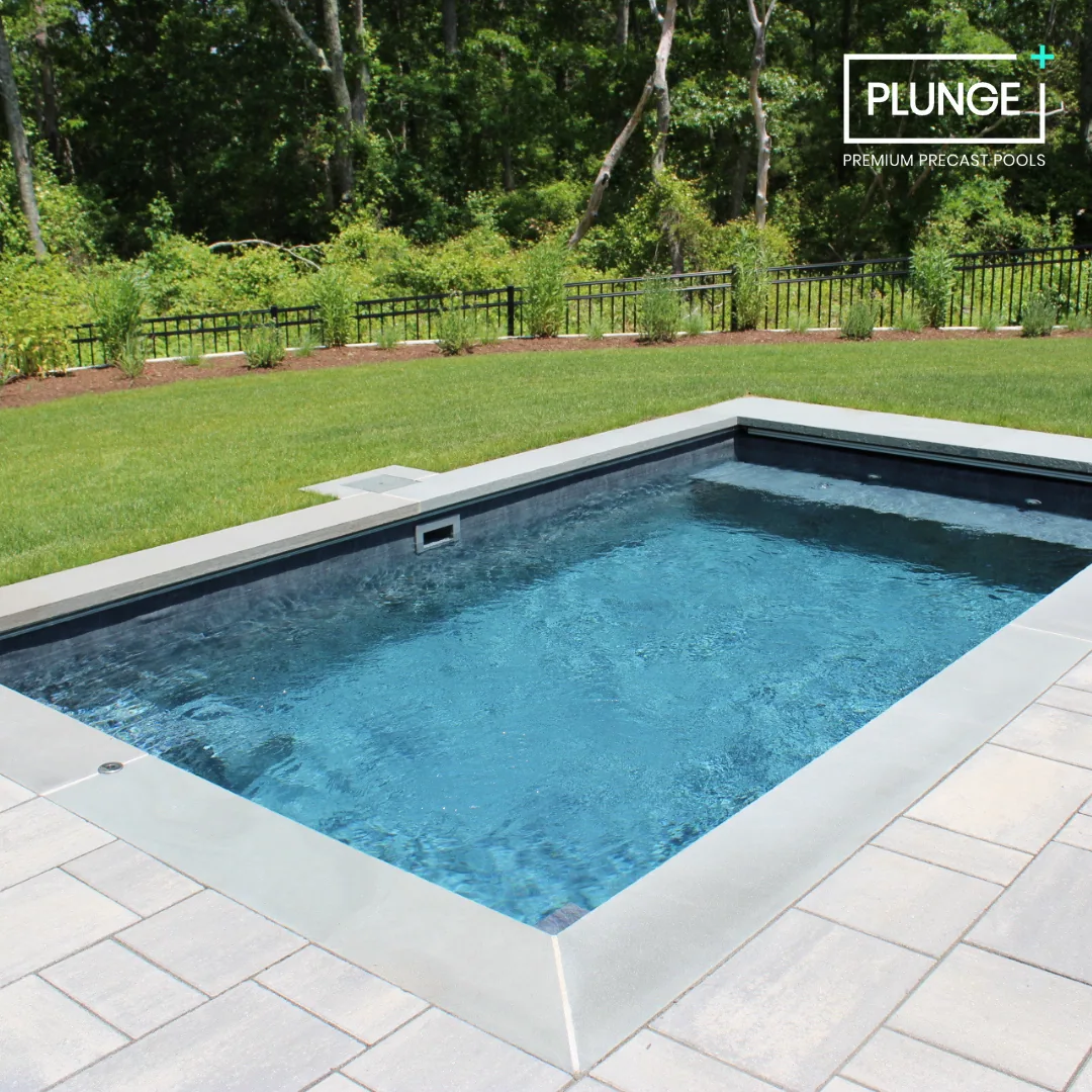 A small rectangular inground pool with clear blue water, surrounded by beige stone tiles, in a backyard with a manicured green lawn and a wooded area in the background.