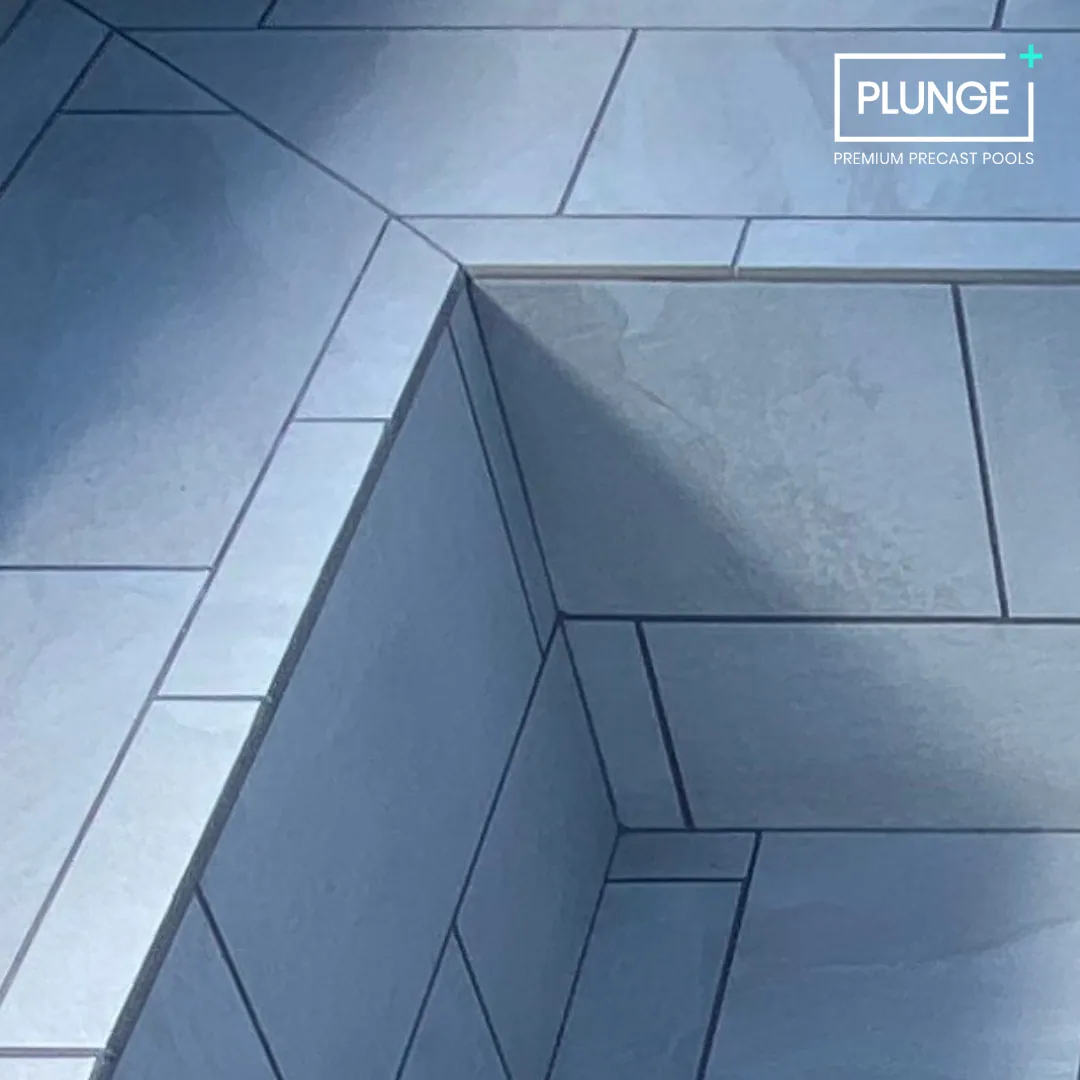 Close-up of a corner of an indoor pool with gray tiles and a step, with a logo in the upper right corner that reads 'PLUNGE Premium Precast Pools.'