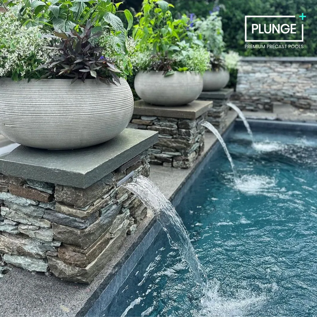 A row of large gray planters with green plants and purple leaves, positioned along a stone waterfall feature at the edge of a pool in a landscaped outdoor space.