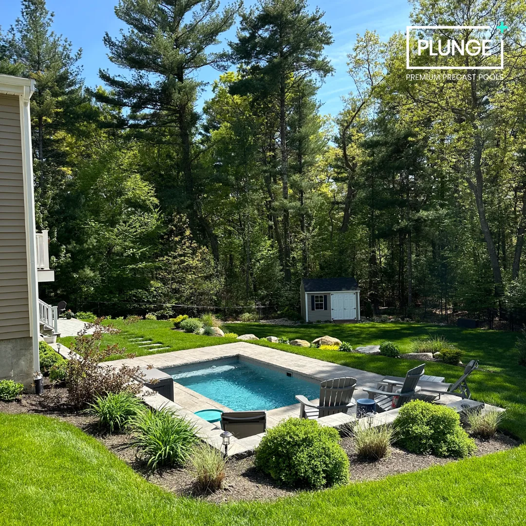 Backyard with a small rectangular swimming pool surrounded by a stone patio, lawn, and garden plants. Lounge chairs are positioned near the pool, with a shed and wooded area in the background. The sky is blue with some clouds.