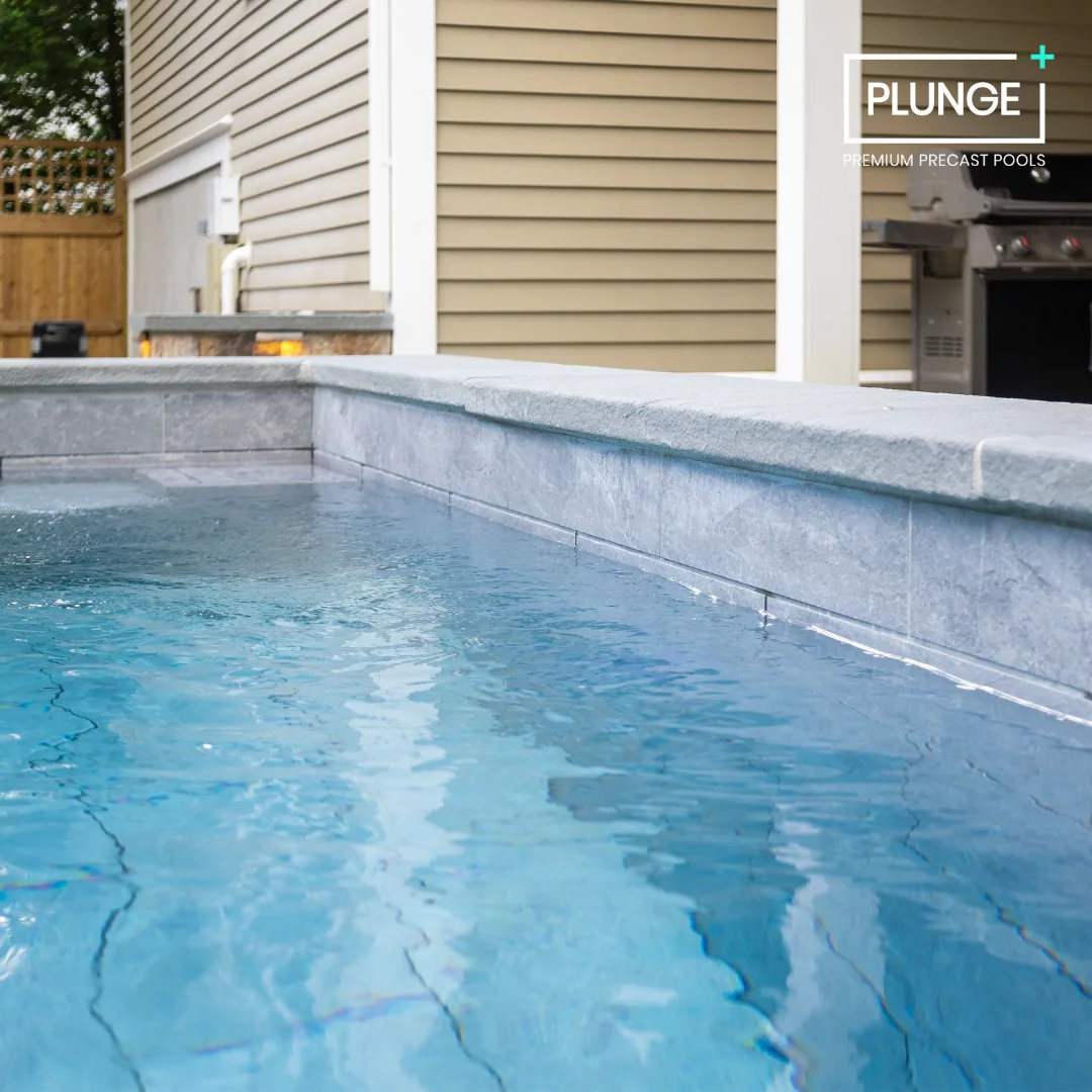 Close-up of a rectangular outdoor swimming pool with gray stone tiles, adjacent to a beige house with vinyl siding, a grill, and a wooden fence in the background.