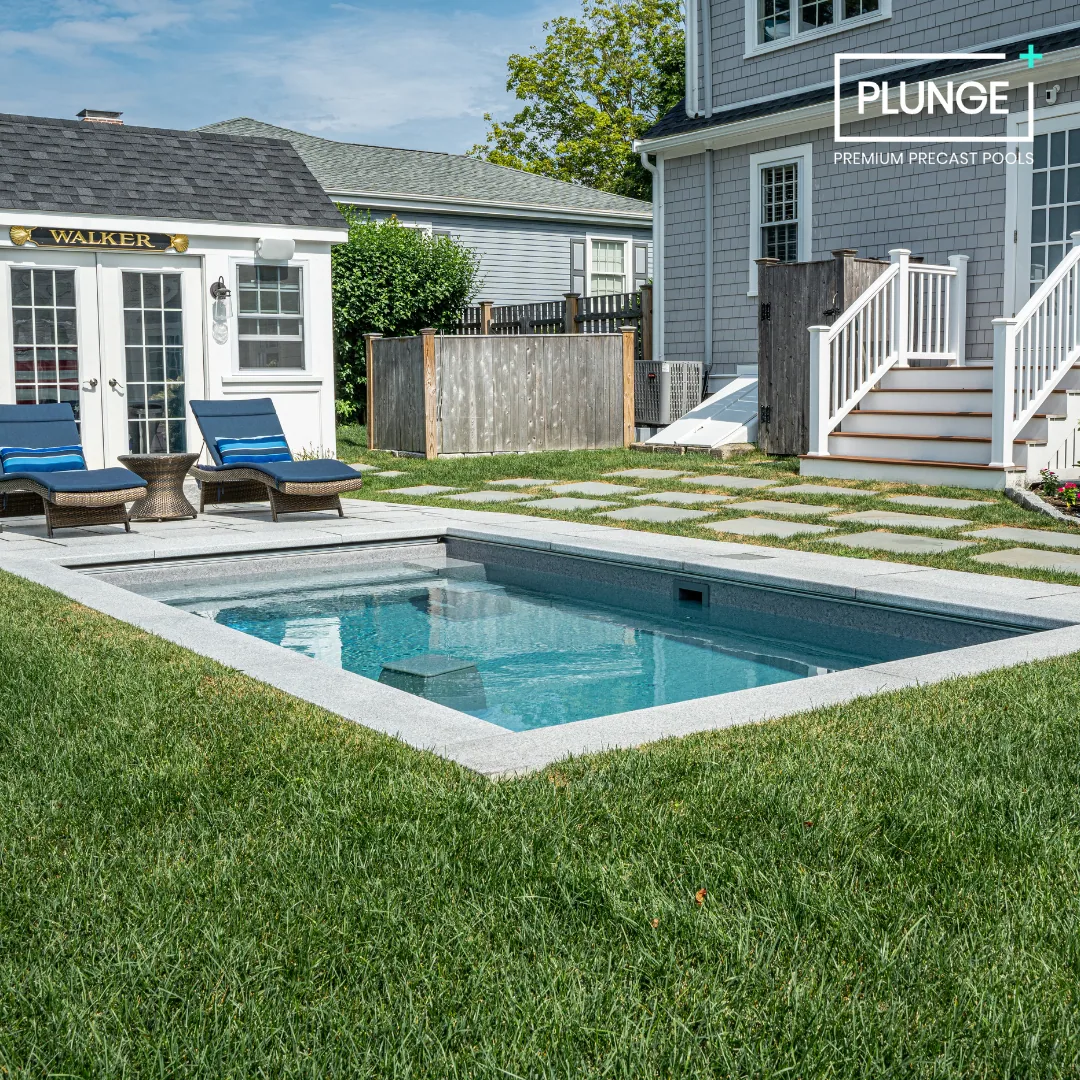 Backyard with a small rectangular swimming pool, two lounge chairs with blue cushions, a side table, a wooden fence, and a house with steps leading to a deck. The scene is bright and sunny.