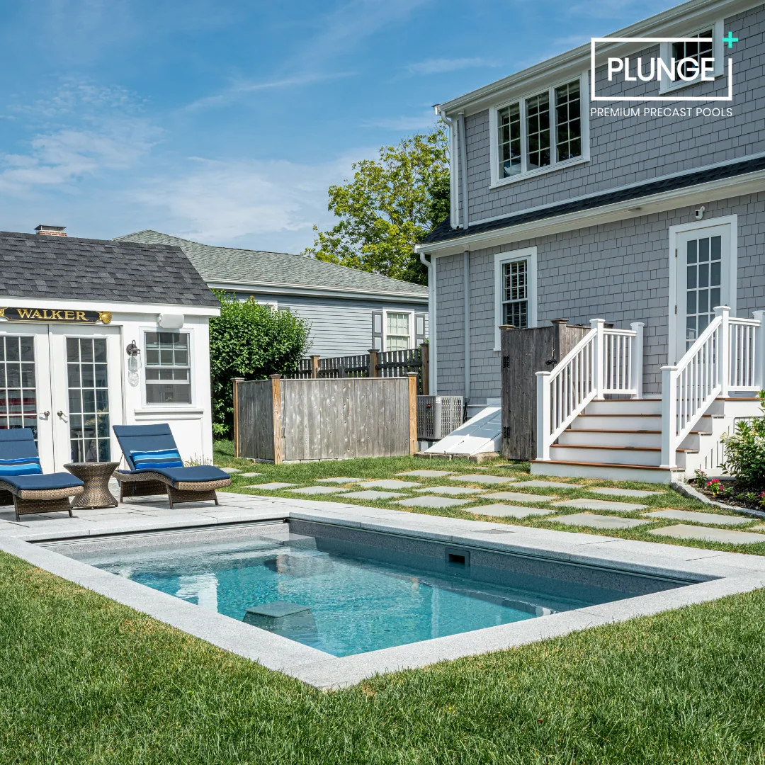 Backyard with a small rectangular pool, two lounge chairs with cushions, a small wicker table, a shed, a wooden fence, and a two-story house with gray siding, white trim, and stairs leading to a door, under a blue sky with some clouds.