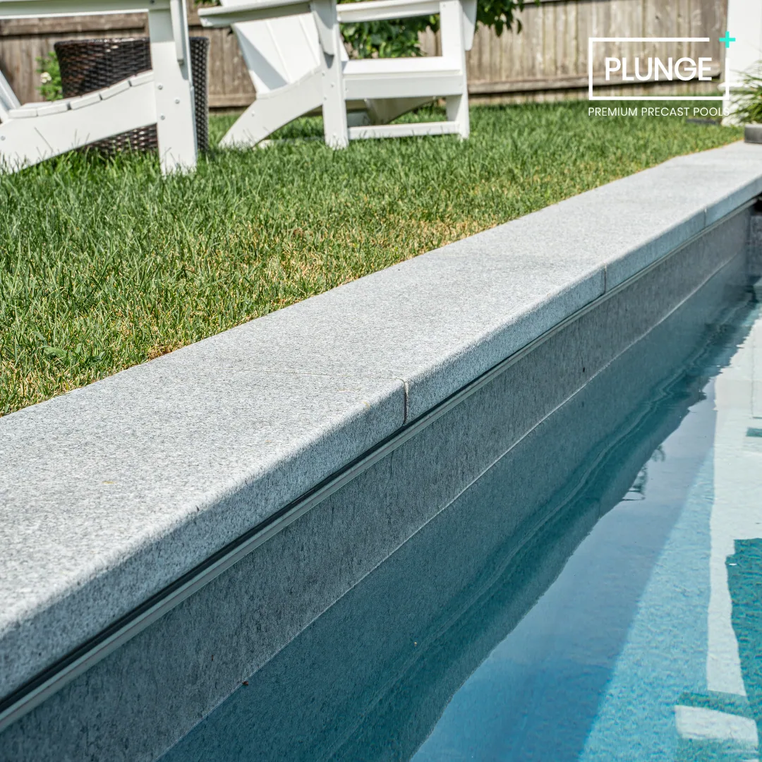 Close-up of a swimming pool with a grey stone edge, adjacent to a grassy yard with outdoor furniture and a wooden fence in the background.