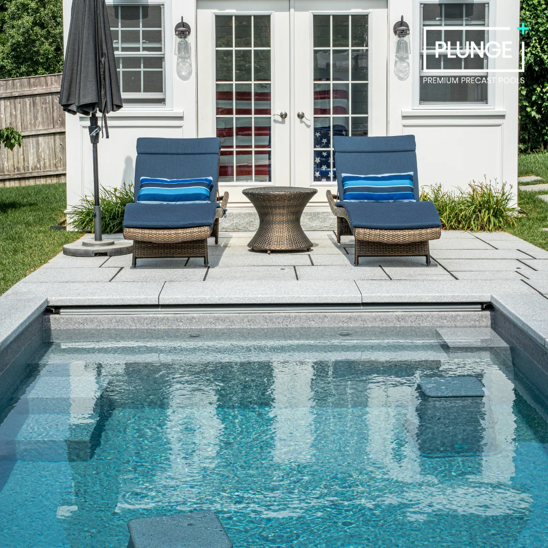 Backyard patio with two blue lounge chairs with striped pillows, a small wicker table between them, an umbrella, and a swimming pool in the foreground, with a house with glass doors behind.