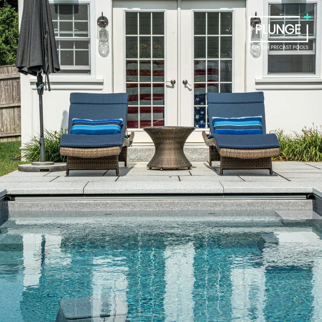 Two blue lounge chairs with pillows, a small round table, an umbrella, and a swimming pool in front of a white house with glass double doors.