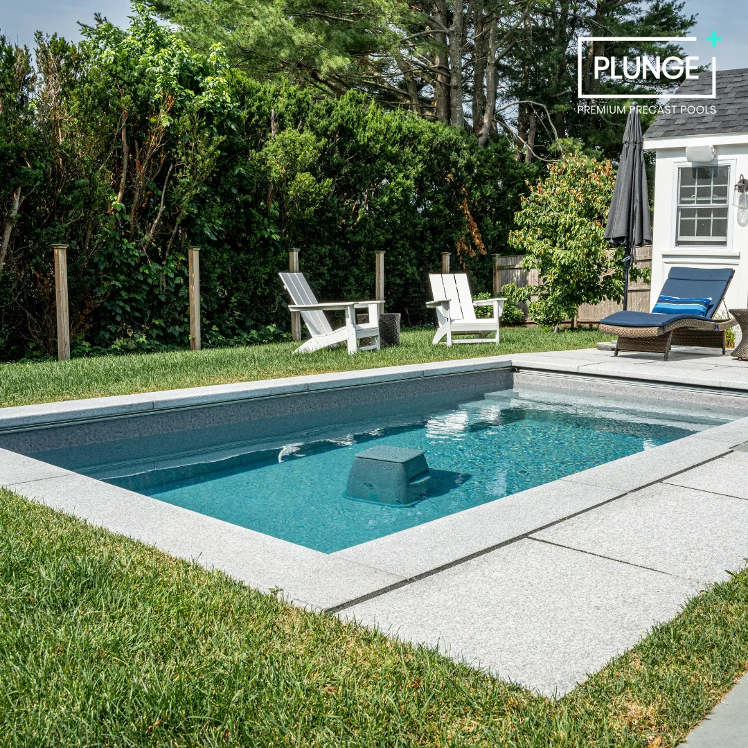 A backyard with a small in-ground pool surrounded by concrete paving and green grass. There are two white Adirondack chairs and a wicker lounge chair with a blue cushion and striped pillow. A black patio umbrella and a small shed or garage are also v
