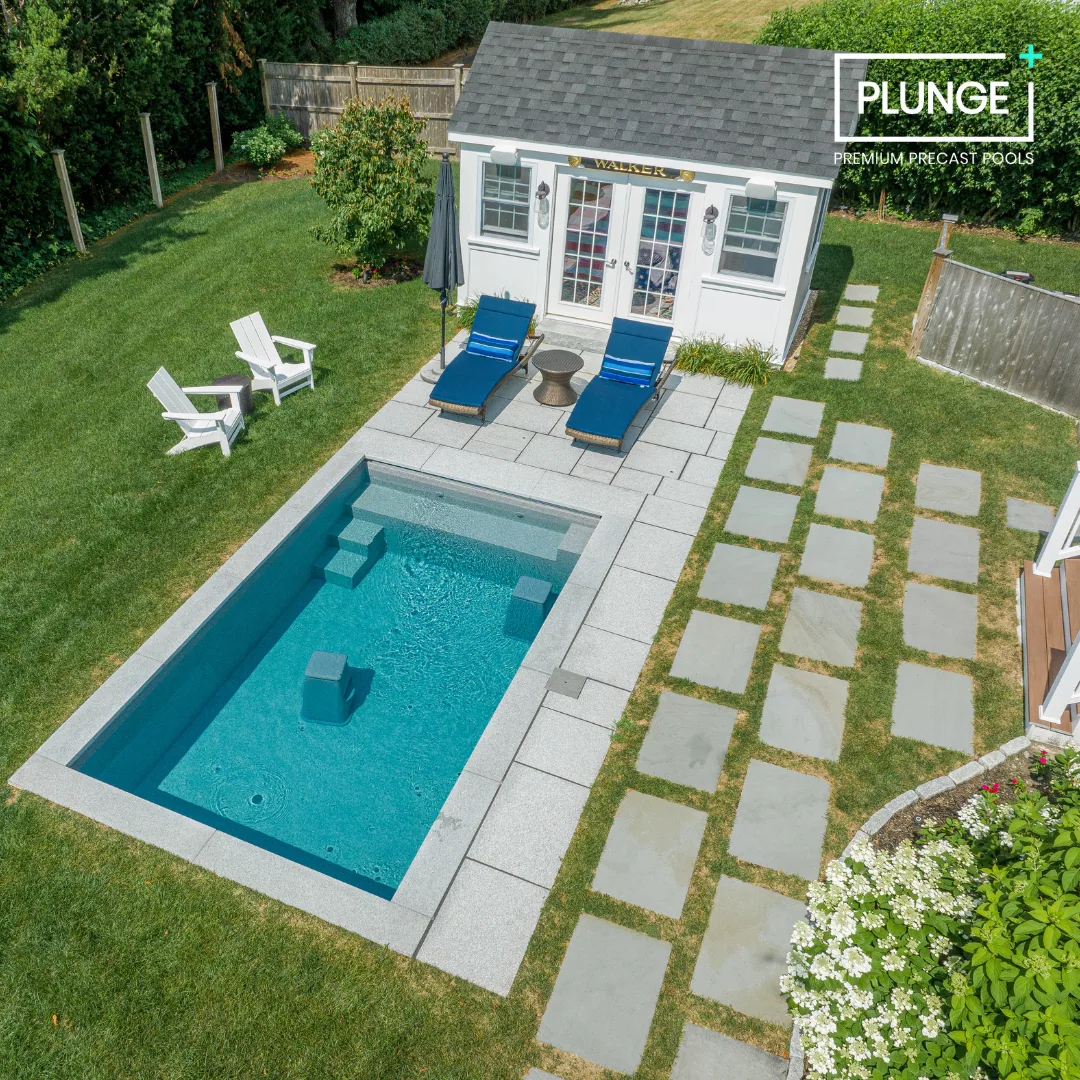 A backyard with a small swimming pool, two blue lounge chairs, a white shed with glass doors, and outdoor seating, surrounded by a green lawn and a wooden fence.