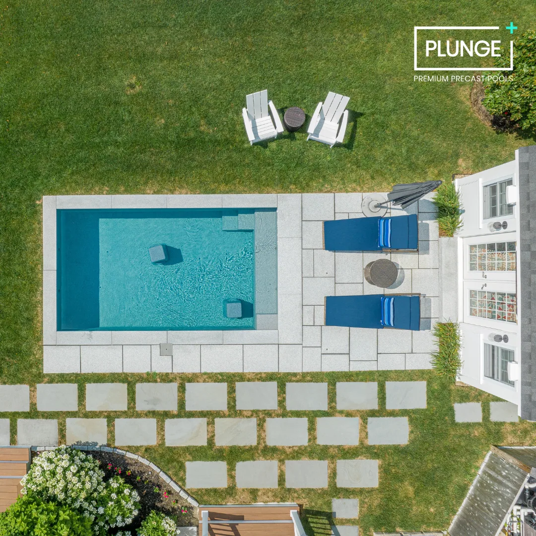A backyard with a rectangular swimming pool, two blue lounge chairs with striped towels, a small round table, and two white Adirondack chairs with a small table on a grassy area. There are stepping stones leading from the house to the pool area.