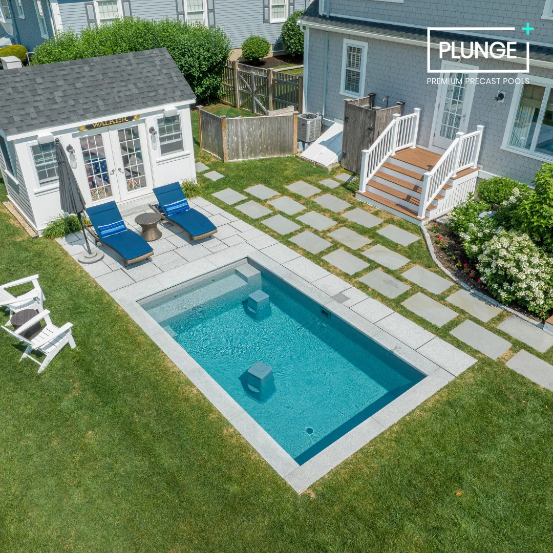 A backyard with a small rectangular swimming pool, two blue lounge chairs with a small table and umbrella, a white shed with a sign that says "Walker," a paved stone pathway, a small wooden deck with stairs, surrounding greenery, and a fence.