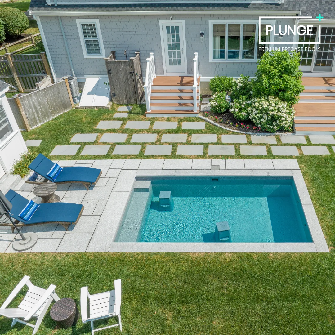 A backyard with a small swimming pool, blue lounge chairs, a small round side table, a white table with chairs, a garden with bushes, a deck with stairs, and a house with a door and windows.