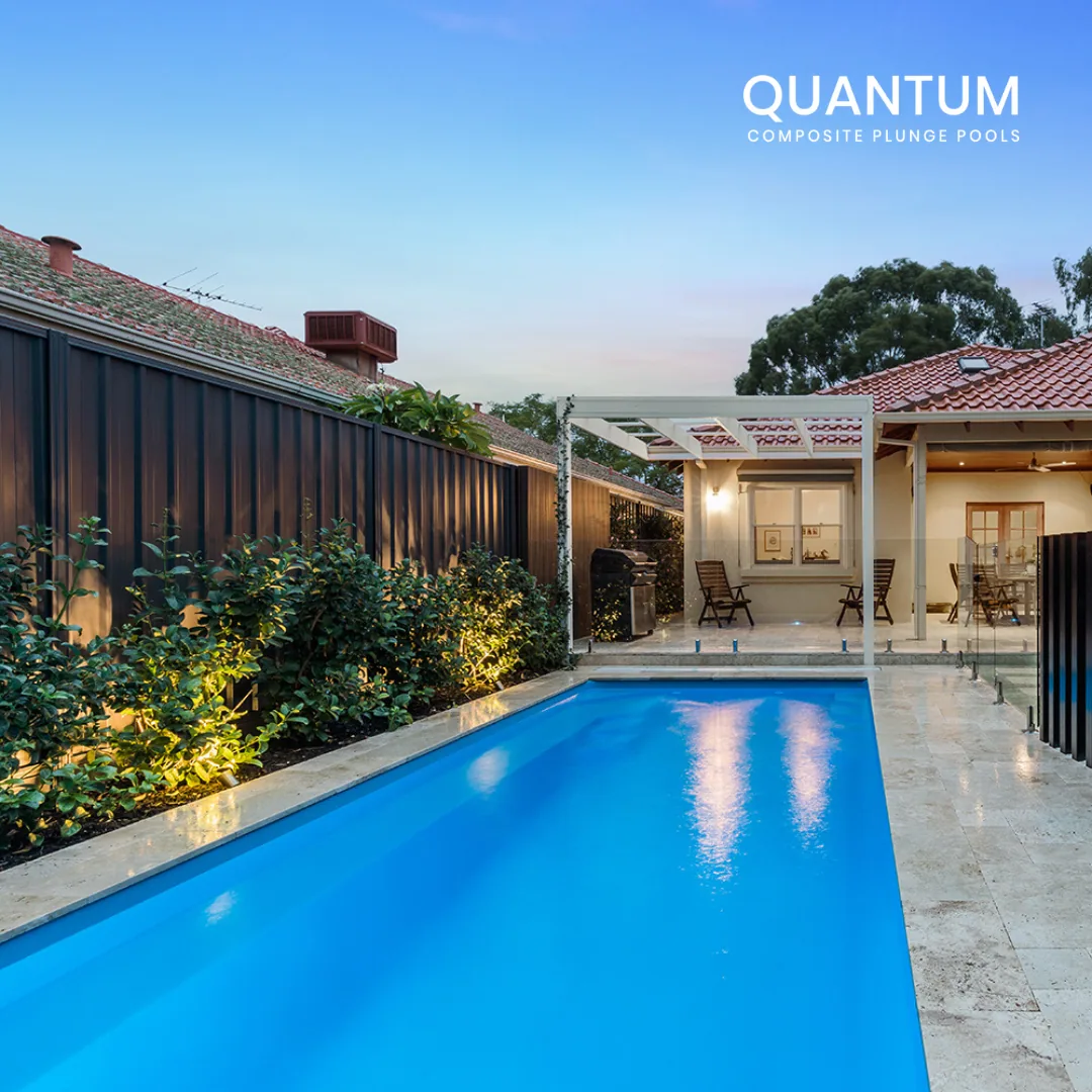 A large beige house with gray roofs, surrounded by green trees. There is a backyard featuring a hot tub, patio furniture, and a deck with outdoor dining. The house has multiple windows, a chimney, and a stairway leading to the yard. The photo is take