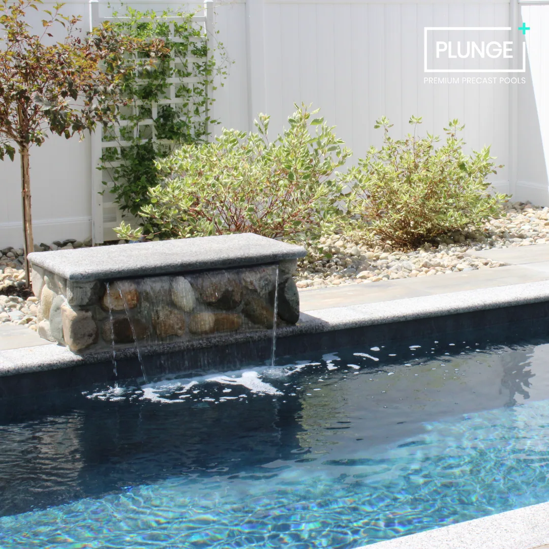 A backyard swimming pool with a stone and concrete waterfall feature, surrounded by landscaped bushes and trees, with a white privacy fence in the background.