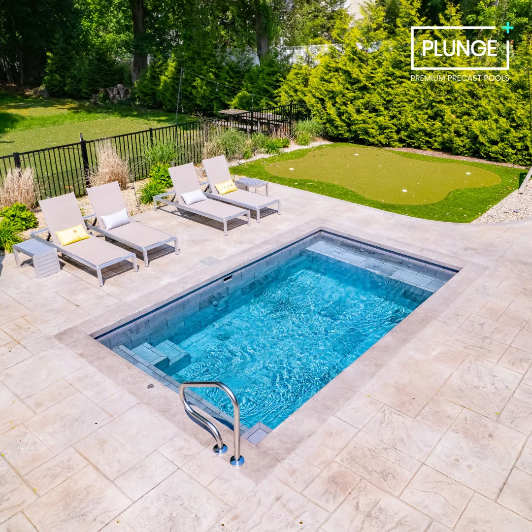 Backyard featuring an in-ground pool with metal handrails, surrounded by a paved deck with four lounge chairs, a small side table, a manicured lawn, and a backdrop of trees and shrubs. Text overlay says 'PLUNGE Premium Precast Pools'.