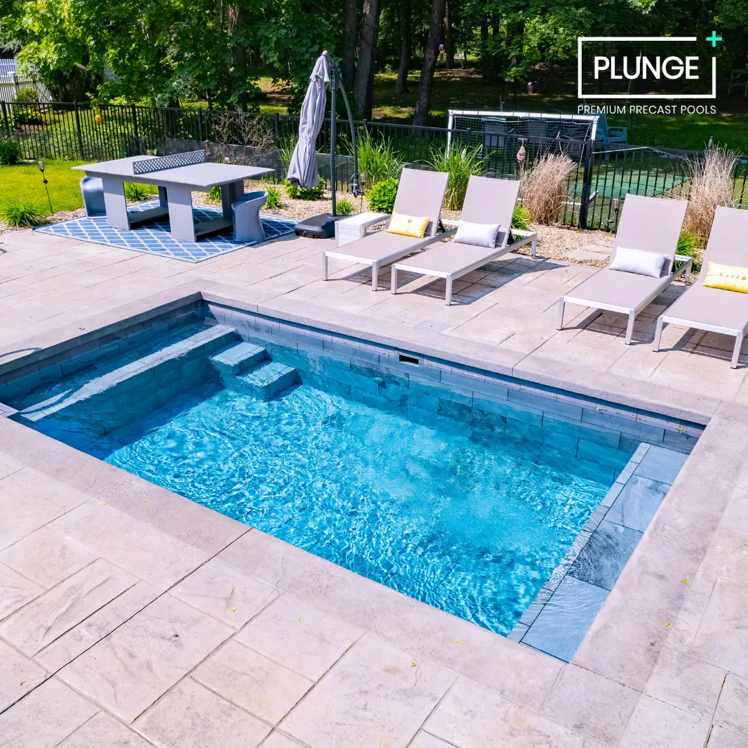 Residential backyard with a rectangular in-ground pool, sun loungers with pillows, a patio table with chairs, and a fenced yard with lush greenery and trees in the background.