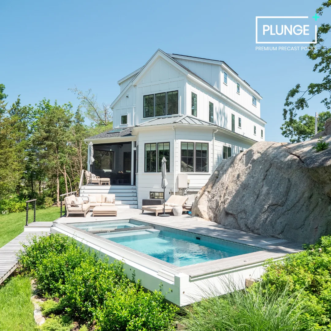 A modern white house with a backyard pool area, surrounded by greenery and a large rock, under a clear blue sky.