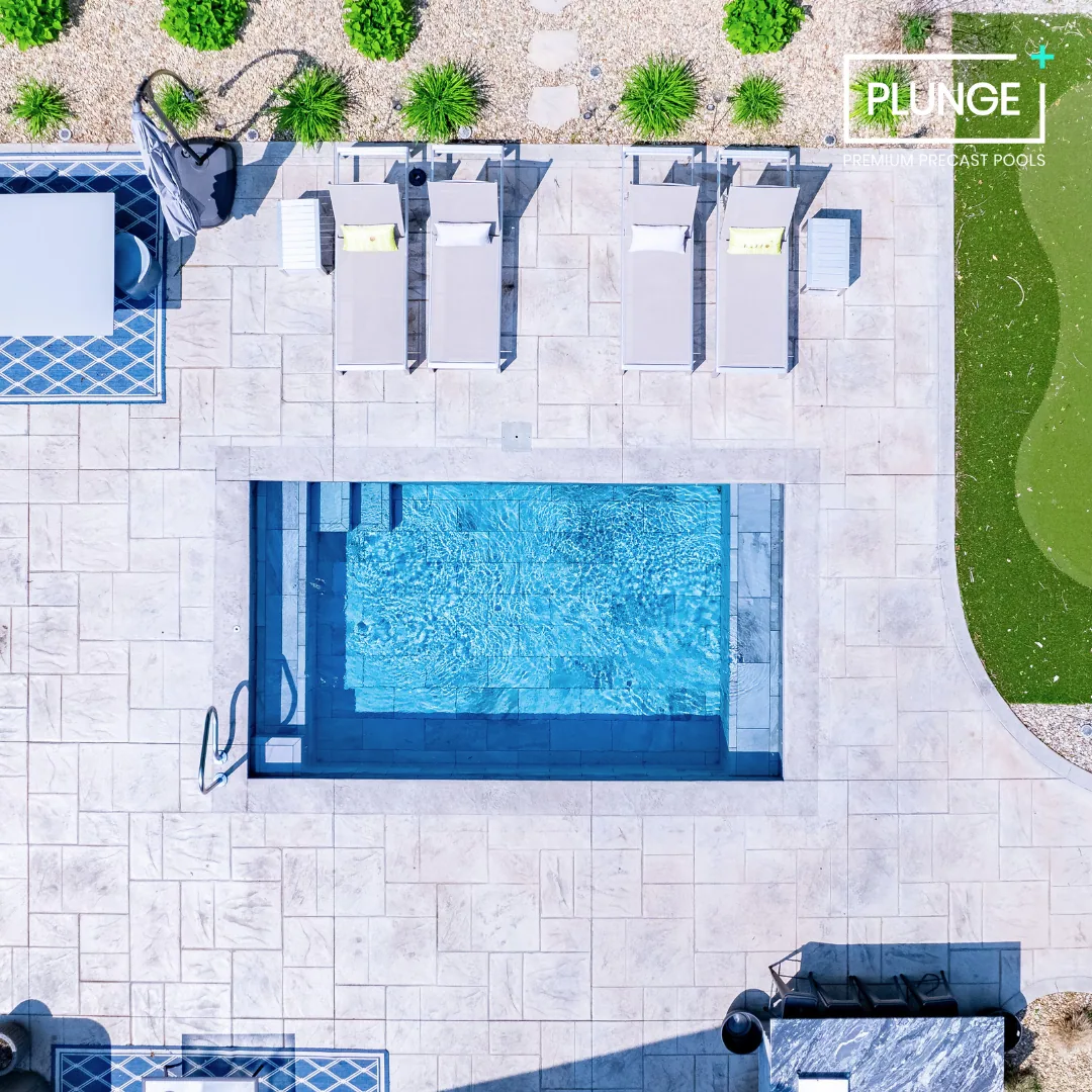 Aerial view of a swimming pool area with four lounge chairs, a small table, and a shaded seating area on a tiled patio, surrounded by greenery and a landscaped yard.