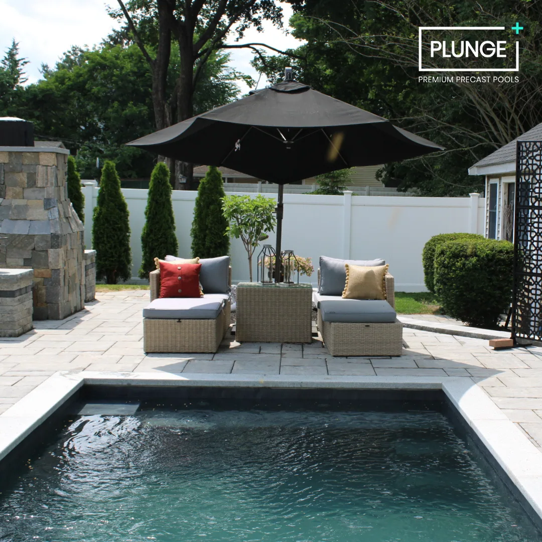Outdoor backyard with a small swimming pool, patio furniture with cushions and pillows, a large umbrella, a stone fireplace, green shrubs, and a white fence in the background.