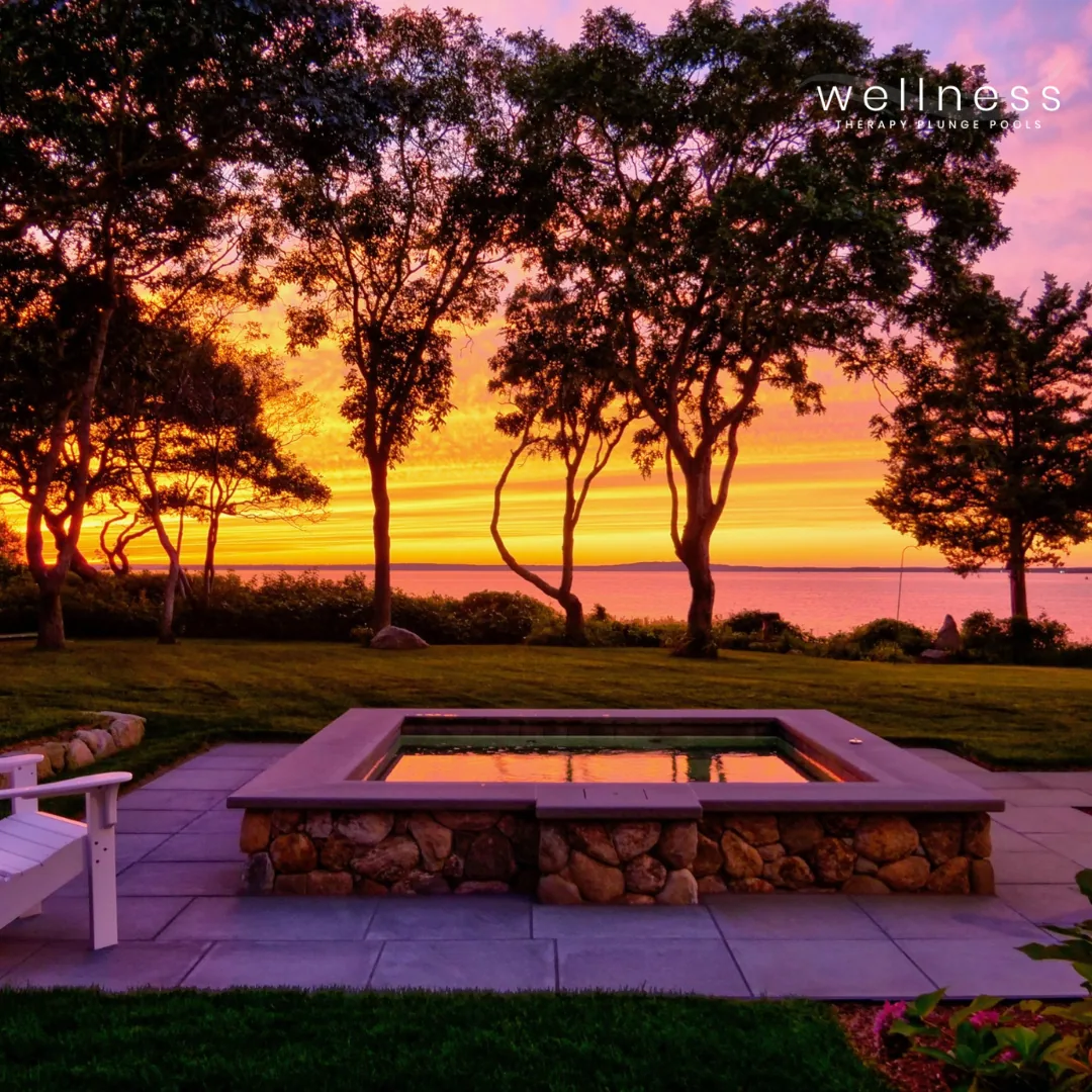 A scenic view of a sunset over a lake, with vibrant orange, pink, and purple skies. In the foreground, there is a hot tub built with stone and wood, surrounded by a patio, with a white bench and lush green grass and trees around.