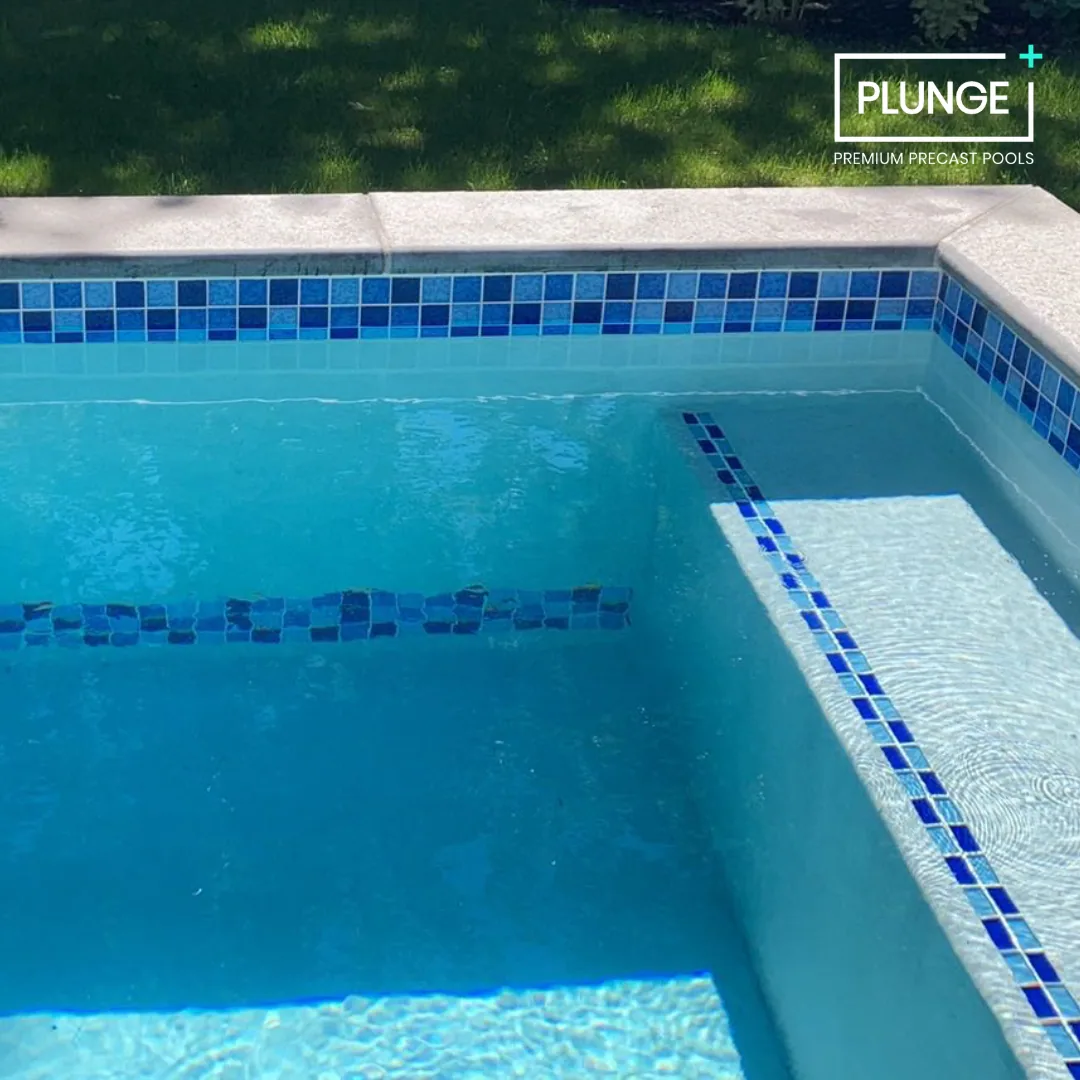 A close-up view of a blue-tiled swimming pool with a small built-in seat, surrounded by a white concrete deck and green grass. The pool has the logo 'PLUNGE' and the words 'Premium Precast Pools' in the top right corner.
