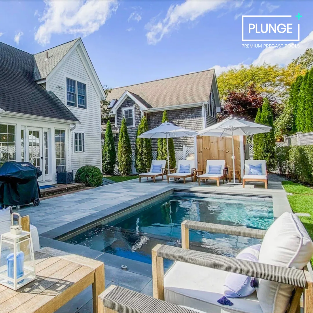 Backyard with a swimming pool, patio chairs with cushions, umbrellas, and a house with white siding and a shingled roof under a partly cloudy sky.