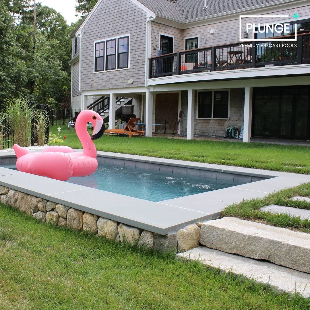 A backyard with a small rectangular swimming pool featuring a pink inflatable flamingo in the water. The pool is surrounded by a stone border and grass. In the background, there is a house with a deck with outdoor furniture and a wooded area behind t