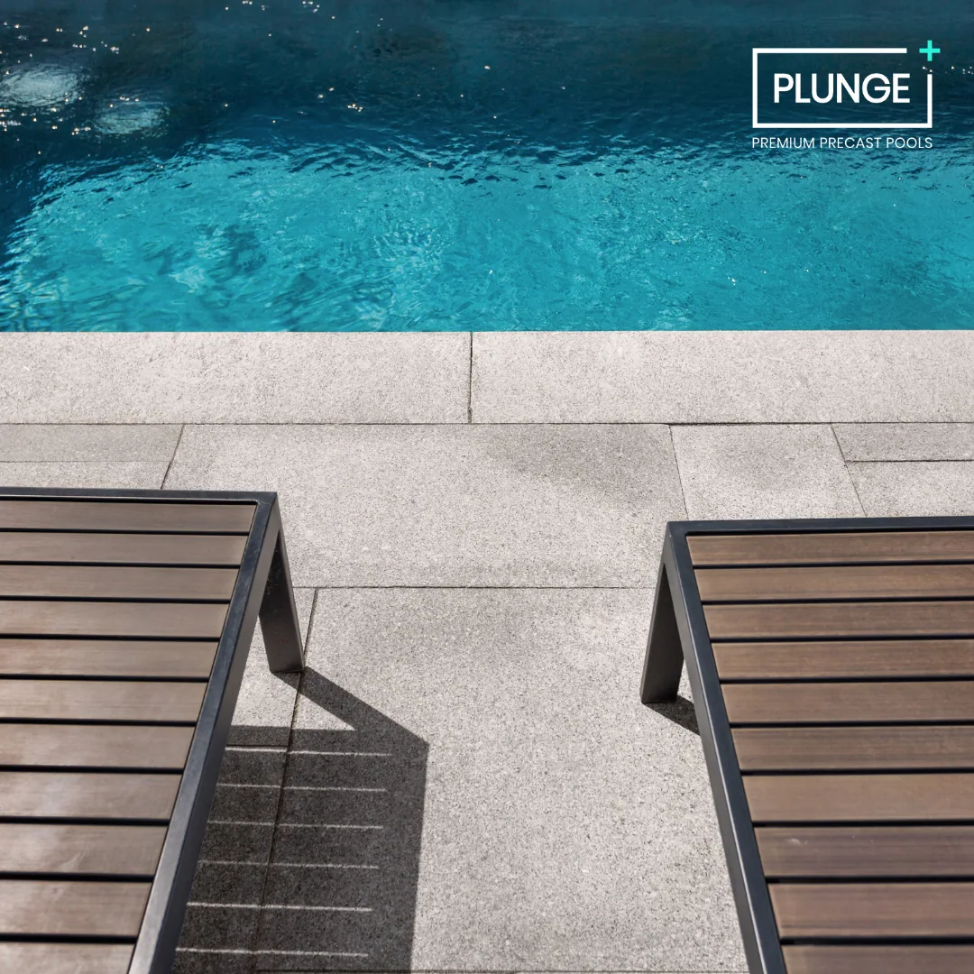 A swimming pool with clear blue water, two lounge chairs on a stone-paved deck, and a logo in the top right corner that reads 'PLUNGE PREMIUM PRECAST POOLS'.