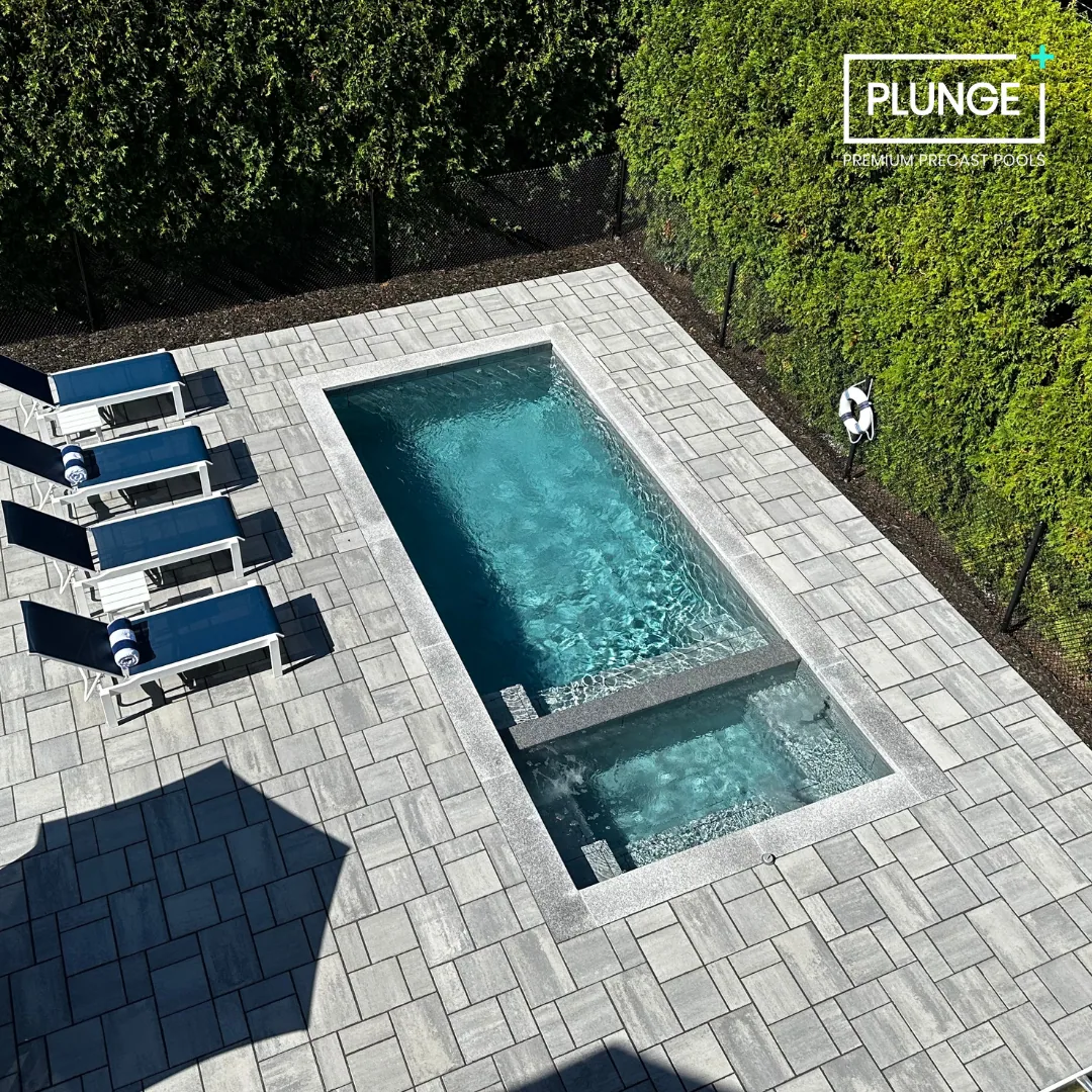 Backyard patio with wicker furniture, a fire pit, a swimming pool, a dining area with a blue umbrella, and garden with trees and bushes behind a white picket fence.