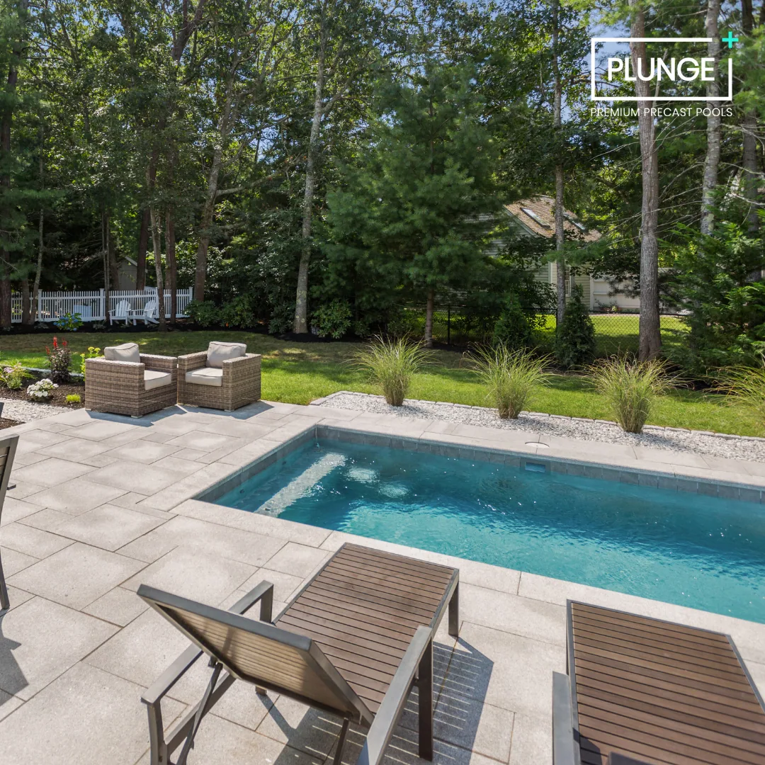 A backyard with a swimming pool, outdoor seating including two lounge chairs, and a small patio area with wicker chairs and a patio table. There are trees and a garden with flowers and ornamental grasses, and a house with a white fence in the backgro