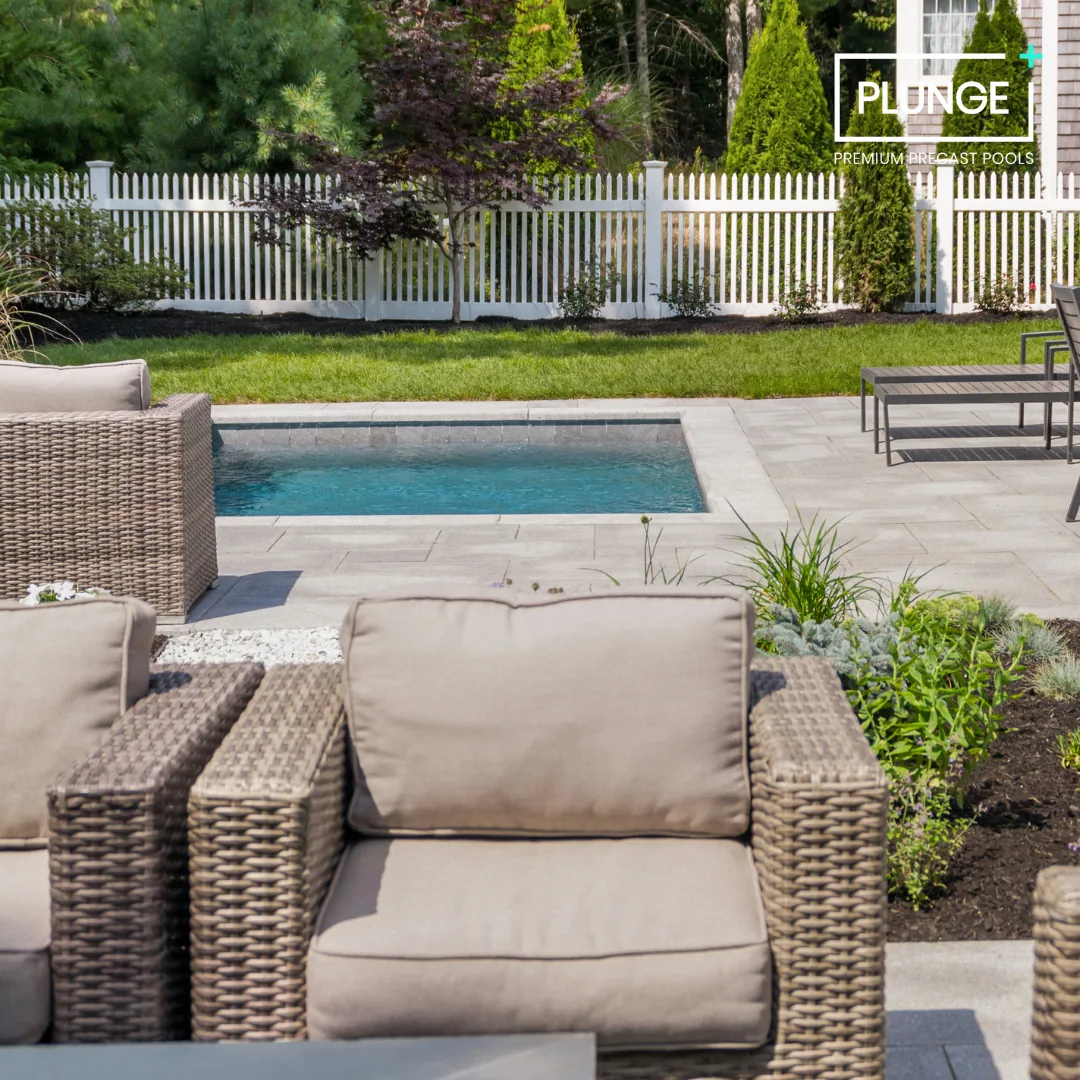 Backyard patio with a small swimming pool, wicker and cushioned outdoor furniture, greenery, and a white picket fence, with a logo and text overlay for Plunge premium precast pools.
