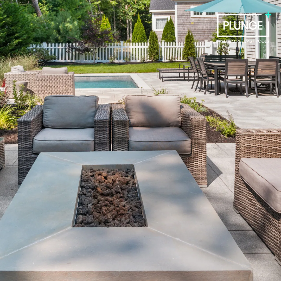 Backyard patio with outdoor furniture, a fire pit, a small swimming pool, and a dining table with chairs under a large umbrella. Green trees and a white picket fence in the background.