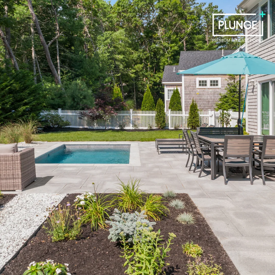 Backyard with a small pool, outdoor table with chairs and a large umbrella, landscaped garden with plants and flowers, white picket fence, and green trees in the background.