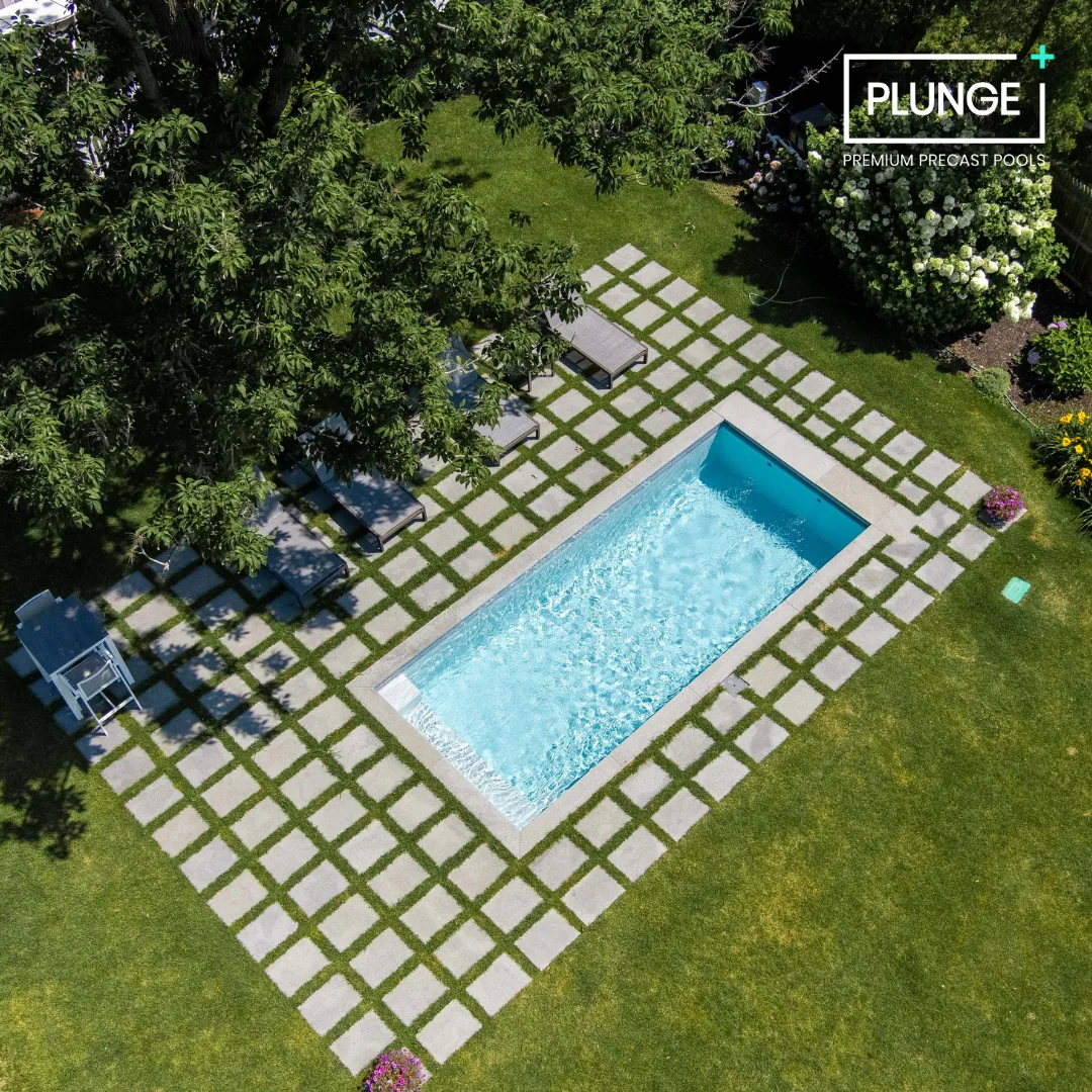 A backyard swimming pool surrounded by a grid pattern of concrete pavers, with several lounge chairs and a table on one side, lush green grass, trees, and flowering bushes.