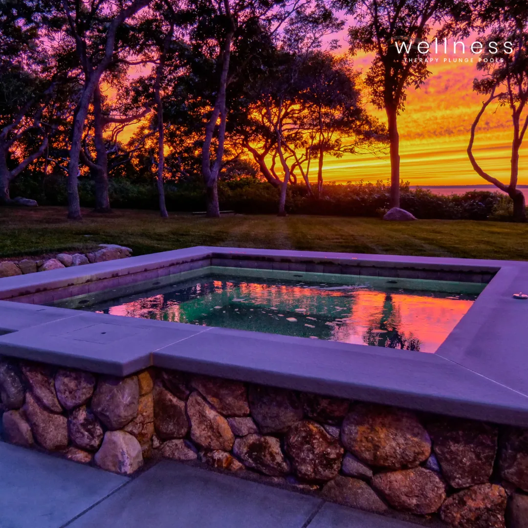 A small hot tub with a stone exterior at sunset, with trees and a colorful sky in the background.
