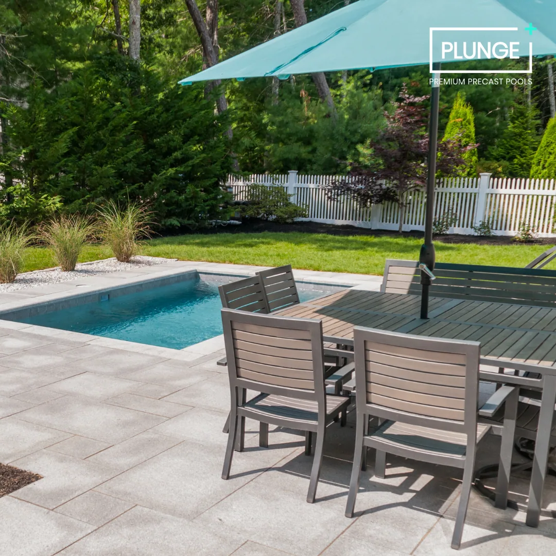 Backyard patio with a small swimming pool, a wooden dining table with four chairs, an umbrella, green grass, and lush trees in the background.