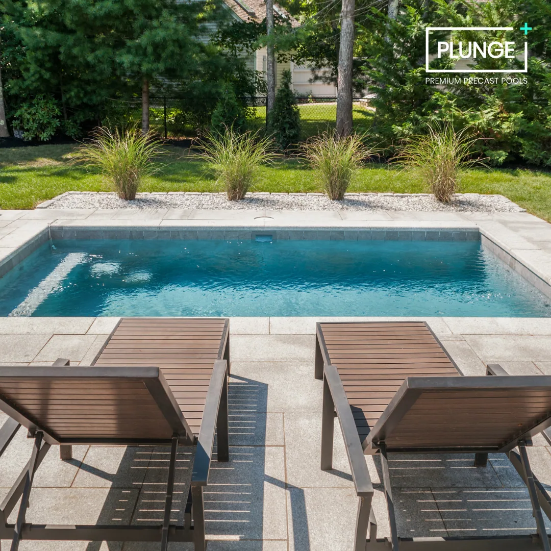 Backyard with a small rectangular pool, two wooden lounge chairs, green grass, and trees in the background. Logo in the top right corner reads 'PLUNGE PREMIUM PRECAST POOLS.'